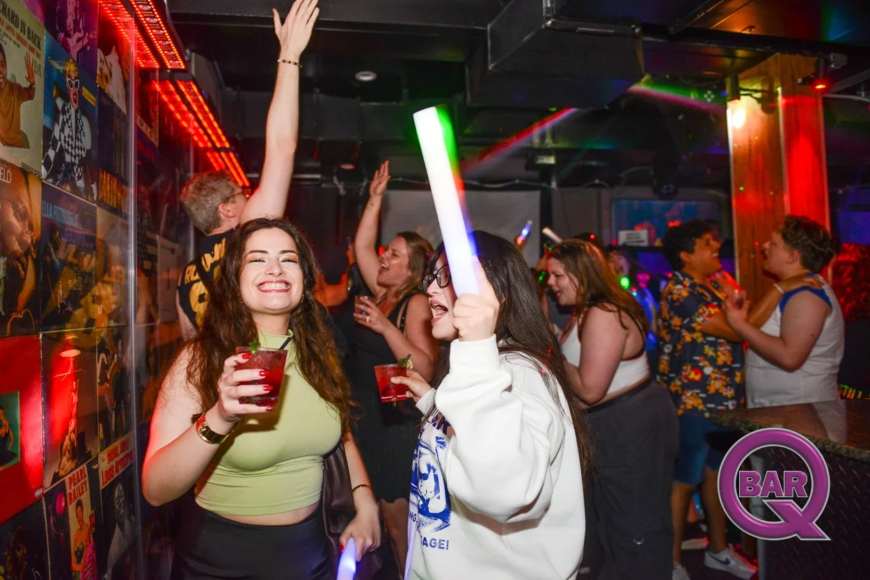 People dancing and celebrating at a nightclub with colorful lights, holding drinks, and enjoying the music. Two women are smiling in the foreground, one with a glow stick and the other with a drink.