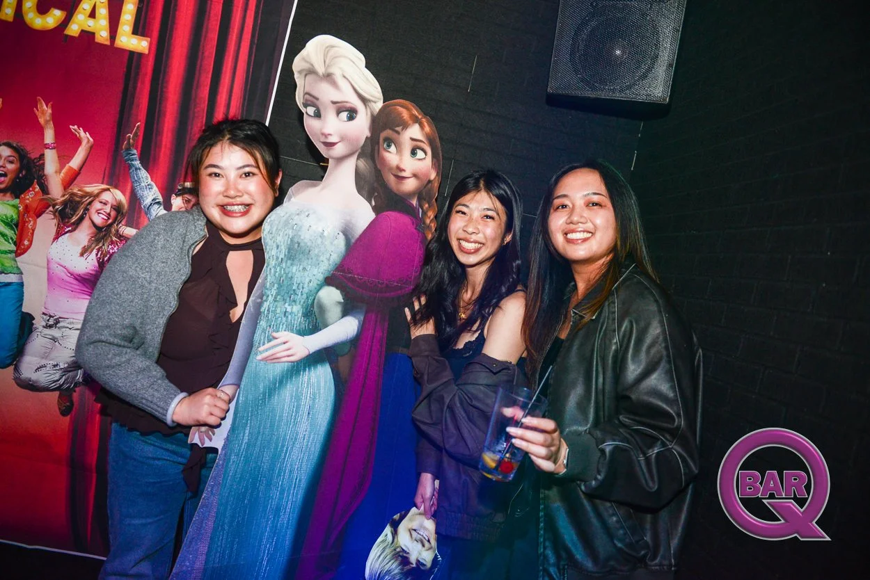 Three young women posing with a life-sized cutout of Elsa and Anna from Frozen in a party setting with a dark wall and a BAR Q logo in the corner.