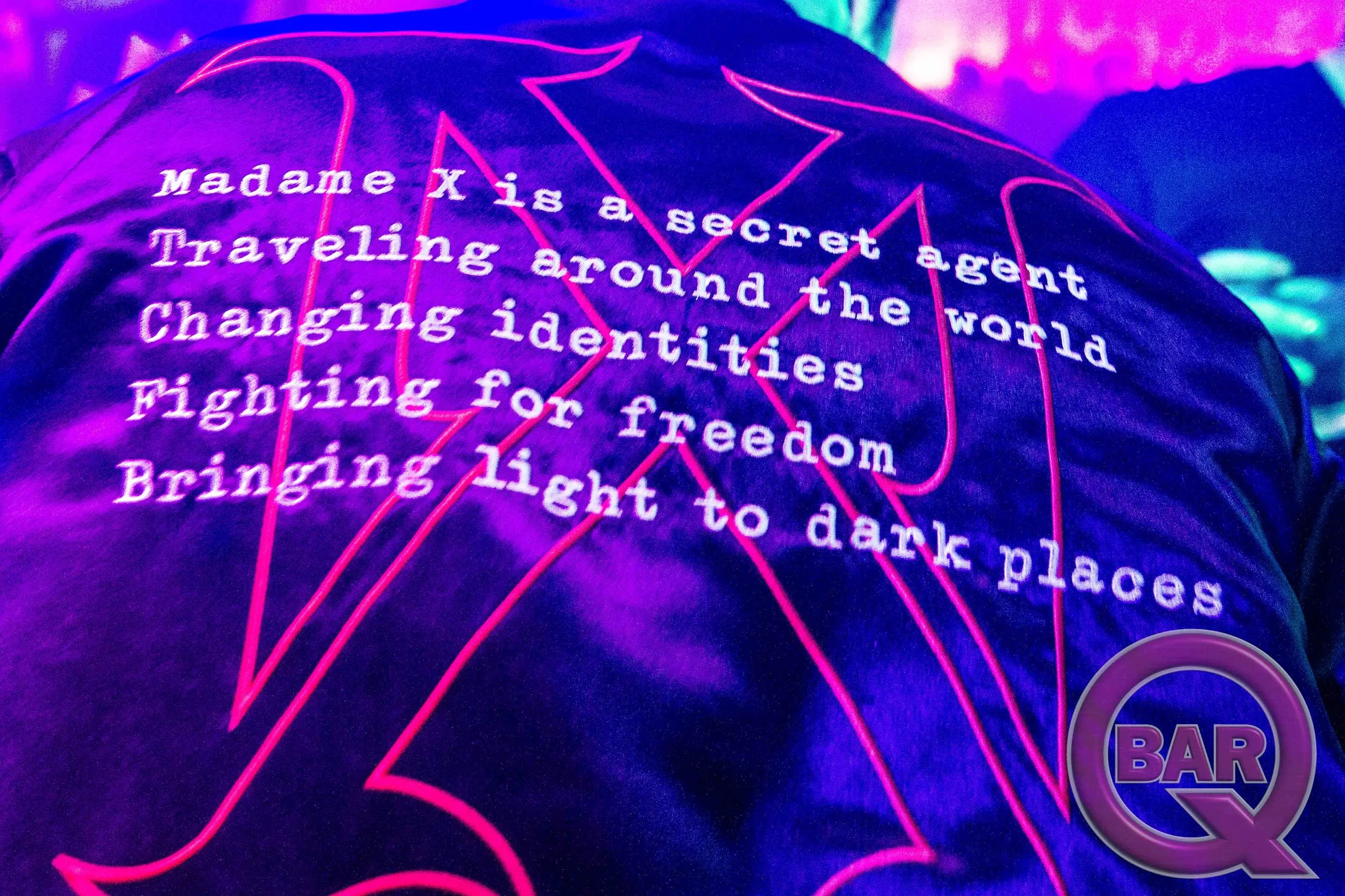Close-up of a neon-lit poster with a star map background, containing the quote 'Madame X is a secret agent traveling around the world changing identities fighting for freedom bringing light to dark places' in white text.