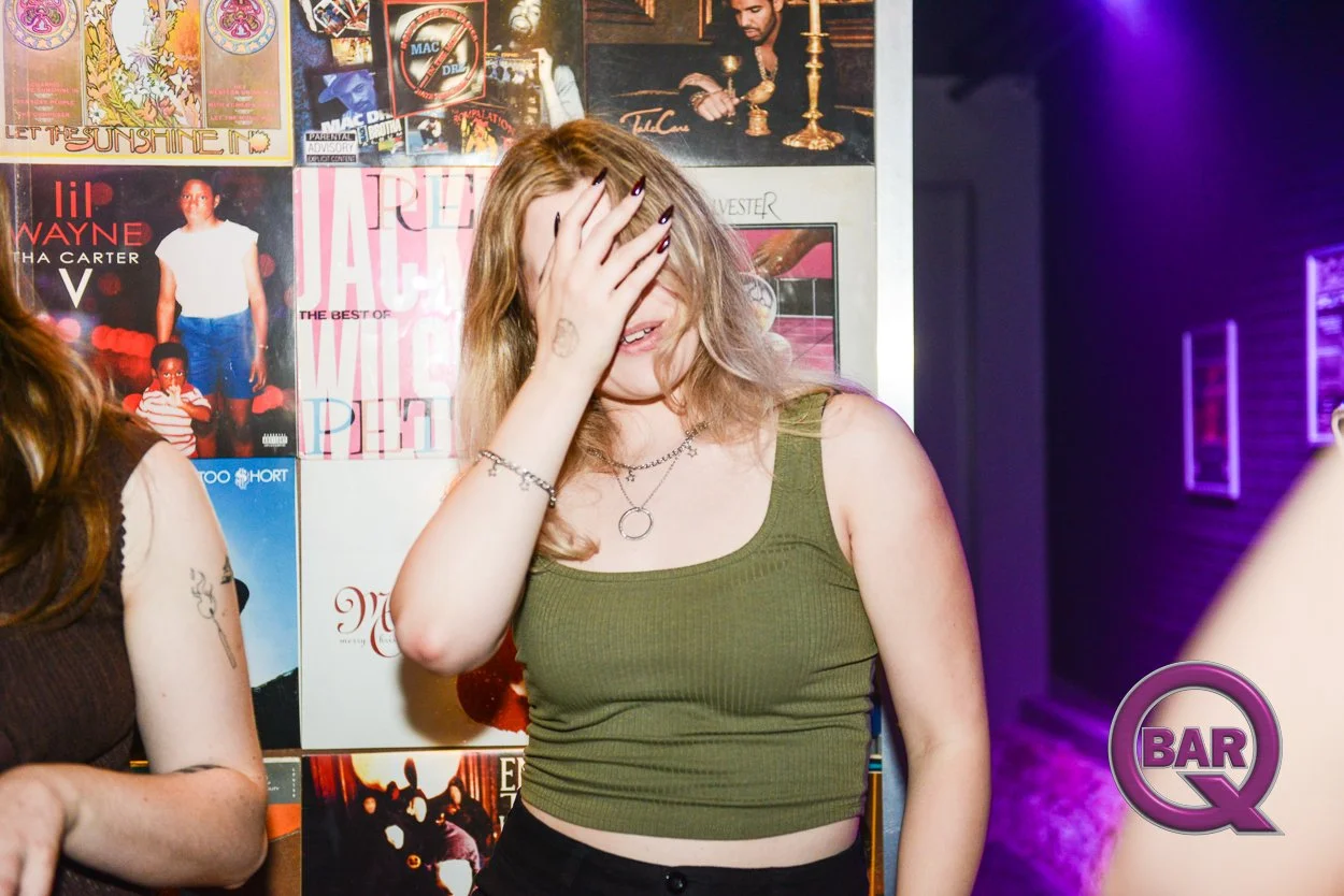 A young woman with light skin and blonde hair, wearing a green tank top and silver jewelry, is covering her face with her hand at a bar or nightclub. The background features posters and album covers on the wall, and the bar logo is visible in the bot