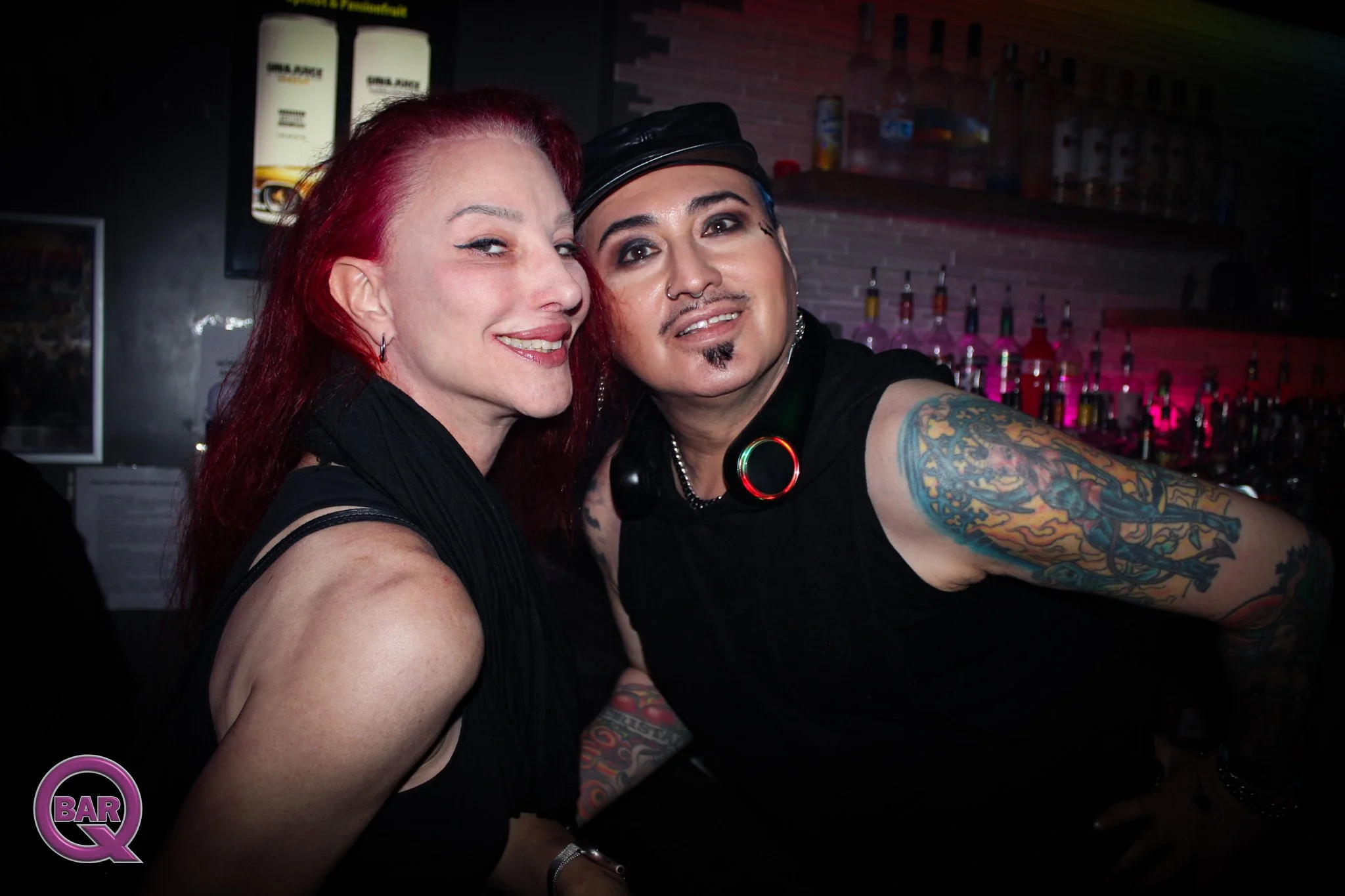 Two friends taking a selfie in a bar, one with long red hair and the other with short dark hair, tattoos, and wearing a black cap, smiling and looking at the camera.
