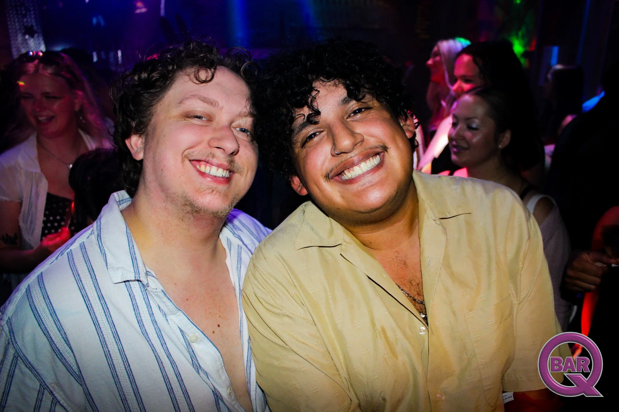 Two smiling men with curly hair and wearing collared shirts, standing close together at a social event or party, with other people in the background.