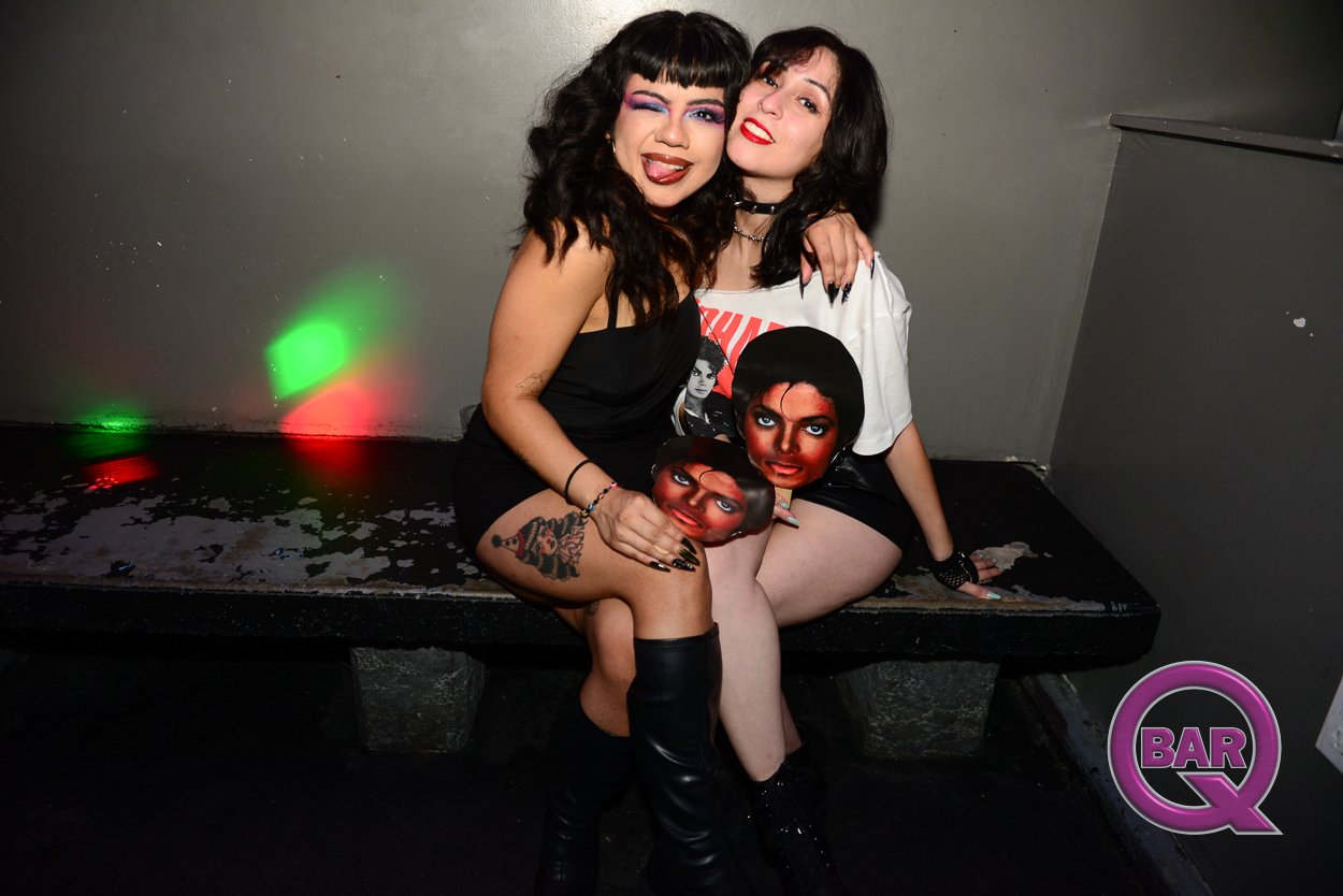 Two women with dark hair and makeup sitting on a bench, embracing and smiling at the camera. One woman has a tattoo on her thigh and is wearing black boots, and the other is wearing a white graphic T-shirt. There are colorful lights and a purple 'Bar