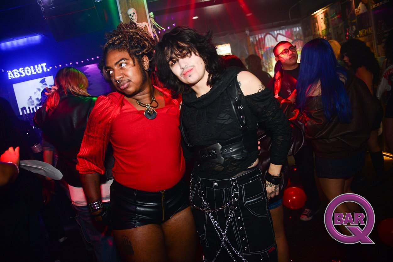 Two individuals with alternative fashion styles posing together in a nightclub. The person on the left has dark skin, dreadlocks, wearing a red top and black leather shorts. The person on the right has light skin, shoulder-length dark hair, wearing a