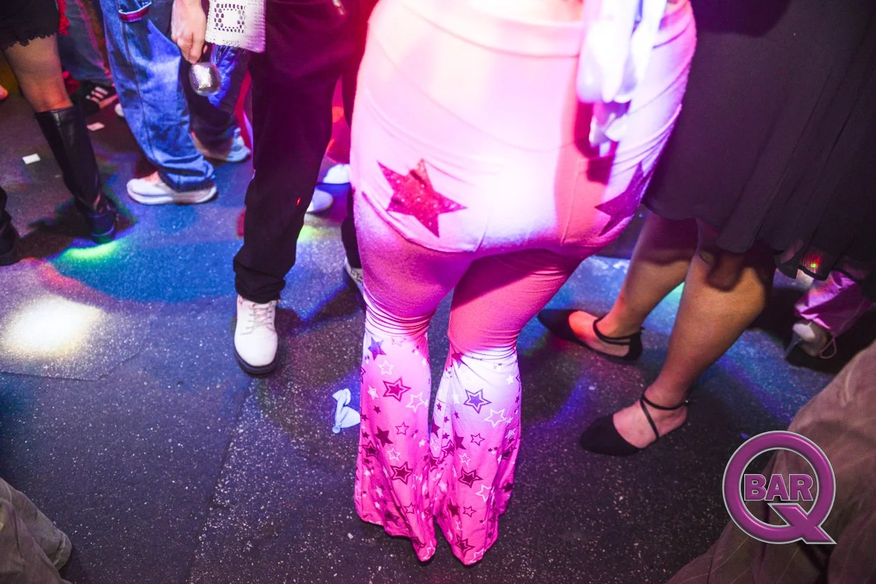 People dancing on a DJ-lit dance floor, focusing on a woman in pink star-patterned flared pants and a white top, with a colorful bar Q logo in the lower right corner.