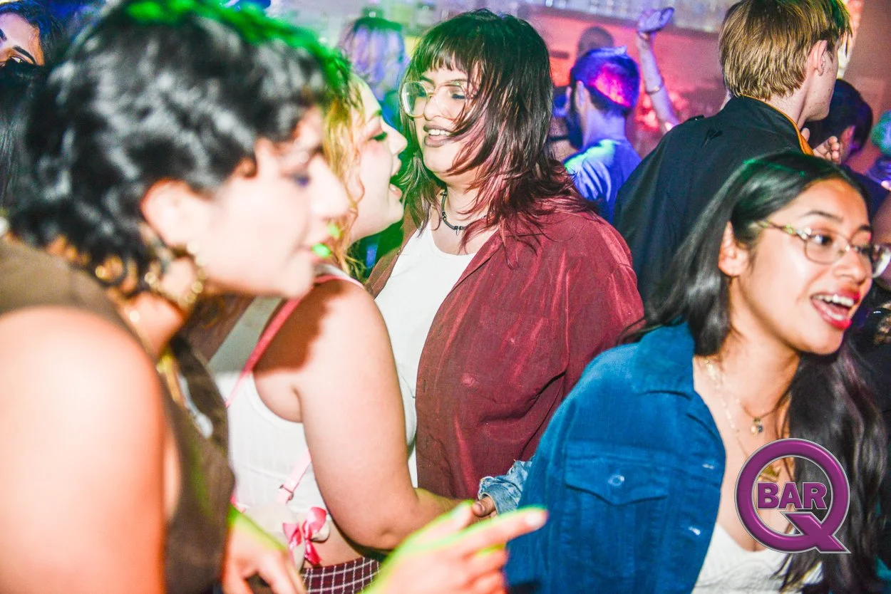 People dancing and socializing at a lively party or nightclub with colorful lighting, including women in stylish clothing and glasses, and a visible 'BAR Q' logo in the bottom right corner.