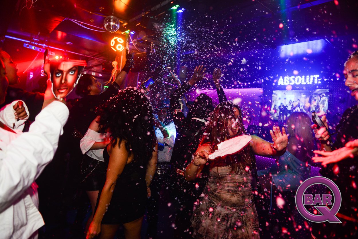 People dancing and celebrating in a nightclub with colorful lights and confetti, with a large screen displaying the word 'ABSOLUT' and a logo reading 'BAR Q' in the corner.