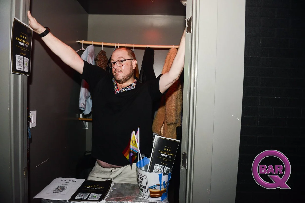 A man standing inside a small room or closet, with arms raised and smiling, wearing glasses and a black shirt with a patterned collar. There are hanging clothes and a coat to his right, and a table with flyers, QR codes, and a flag in front of him. T