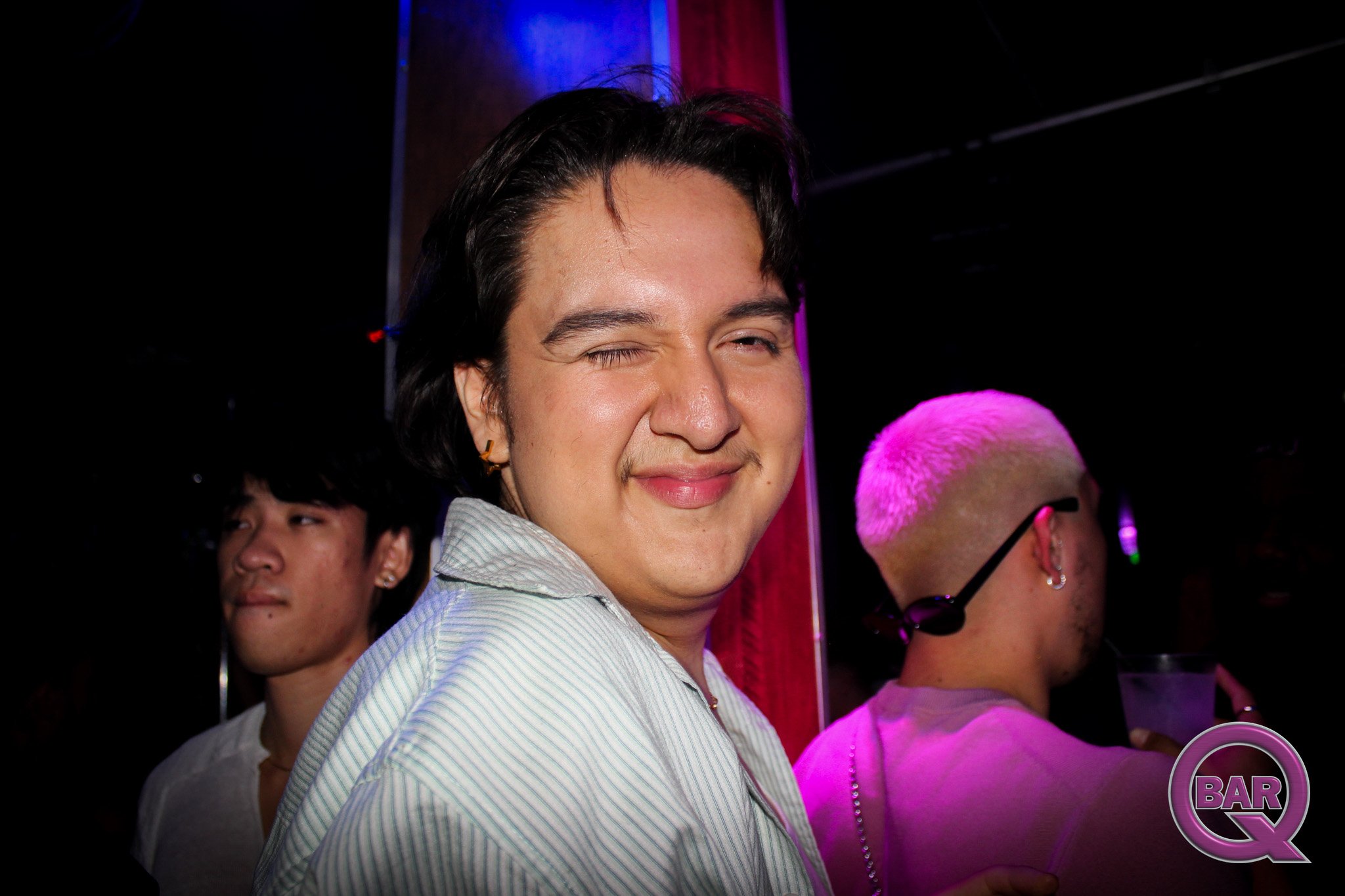 A man with dark hair and a mustache winks at the camera, wearing a striped shirt, surrounded by other people at an indoor event with pink and purple lighting.