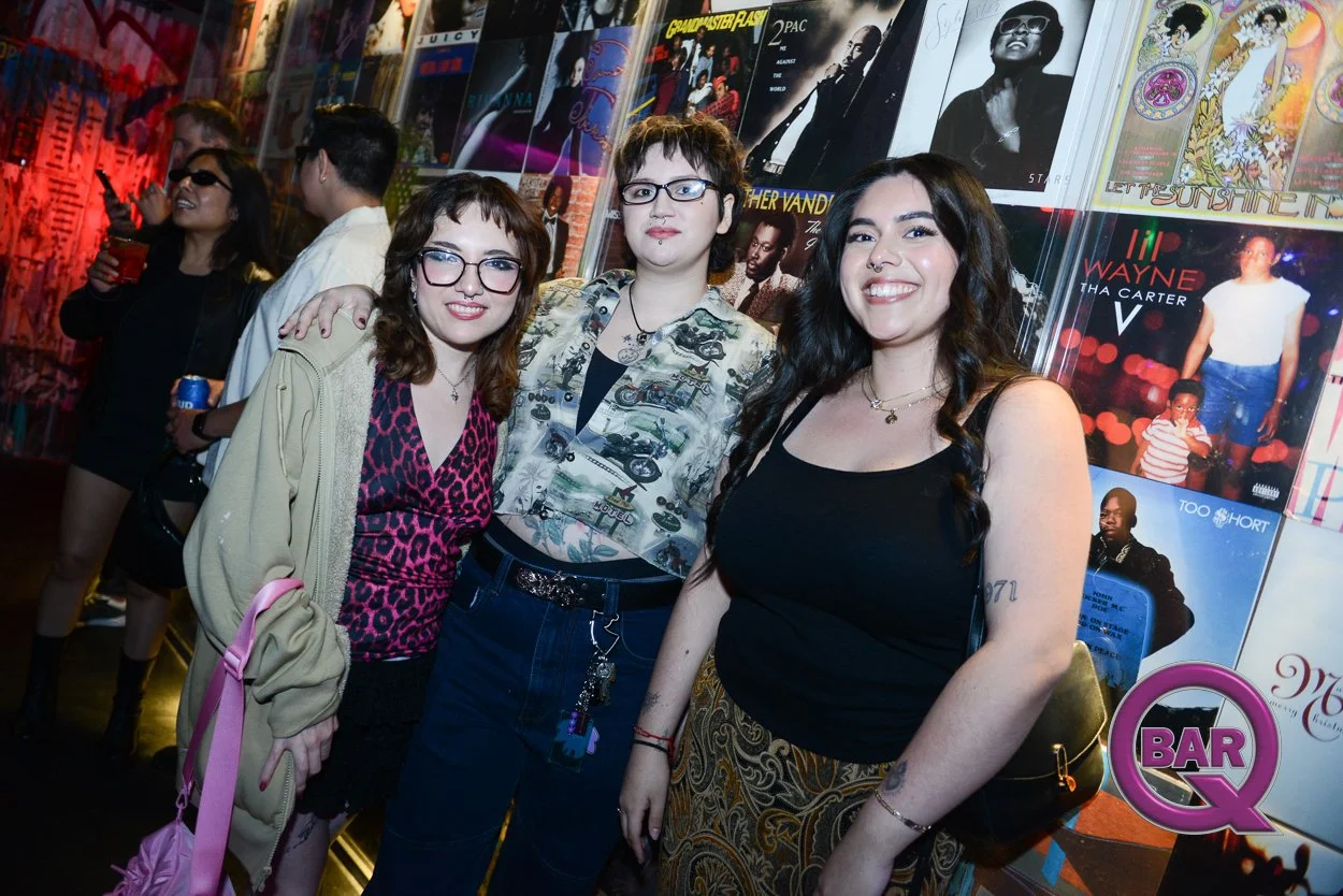 Three women posing and smiling in front of a wall decorated with vinyl record album covers at a music event or bar.