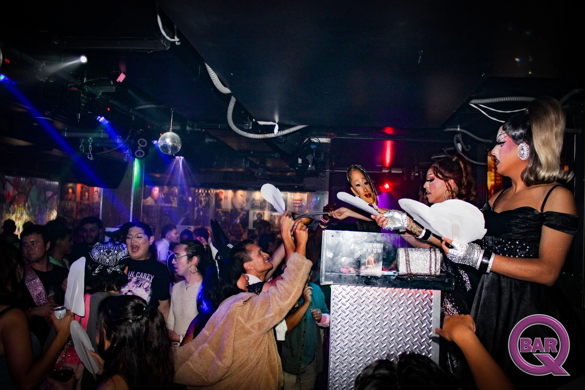 Crowd of enthusiastic people reaching out towards drag queens performing at a bar with neon lights and disco ball in the background.