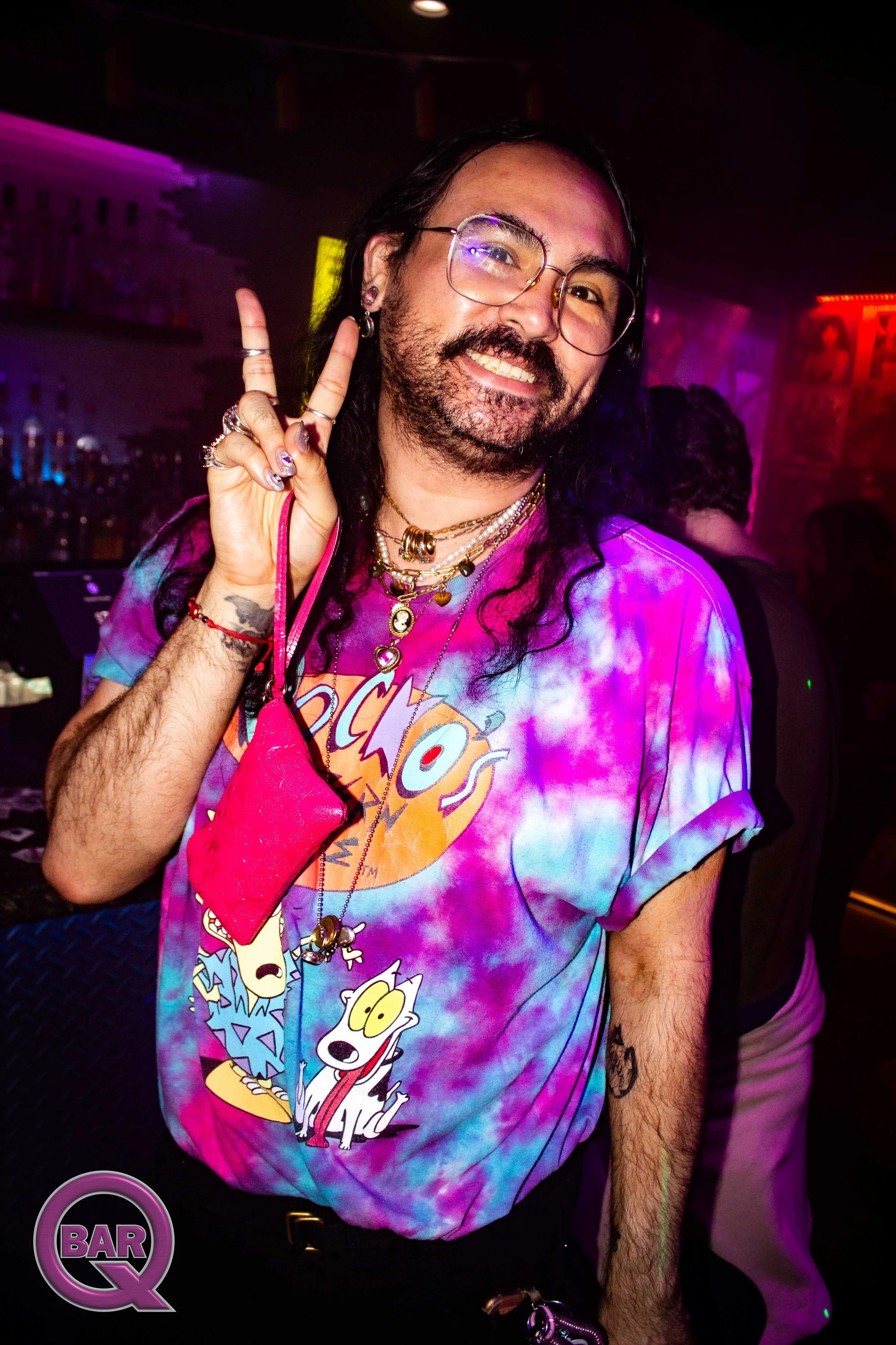 A man with long black hair, beard, and glasses smiling and making a peace sign at a bar with colorful neon lighting. He is wearing a tie-dye shirt featuring cartoon characters.