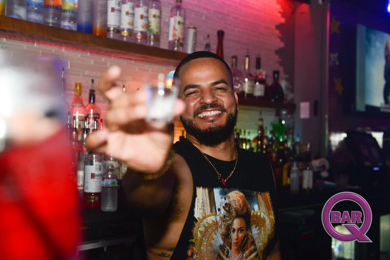 A smiling bartender holding up a shot glass in a bar with colorful bottles on shelves and a television screen in the background, with a purple 'BAR Q' logo in the corner.