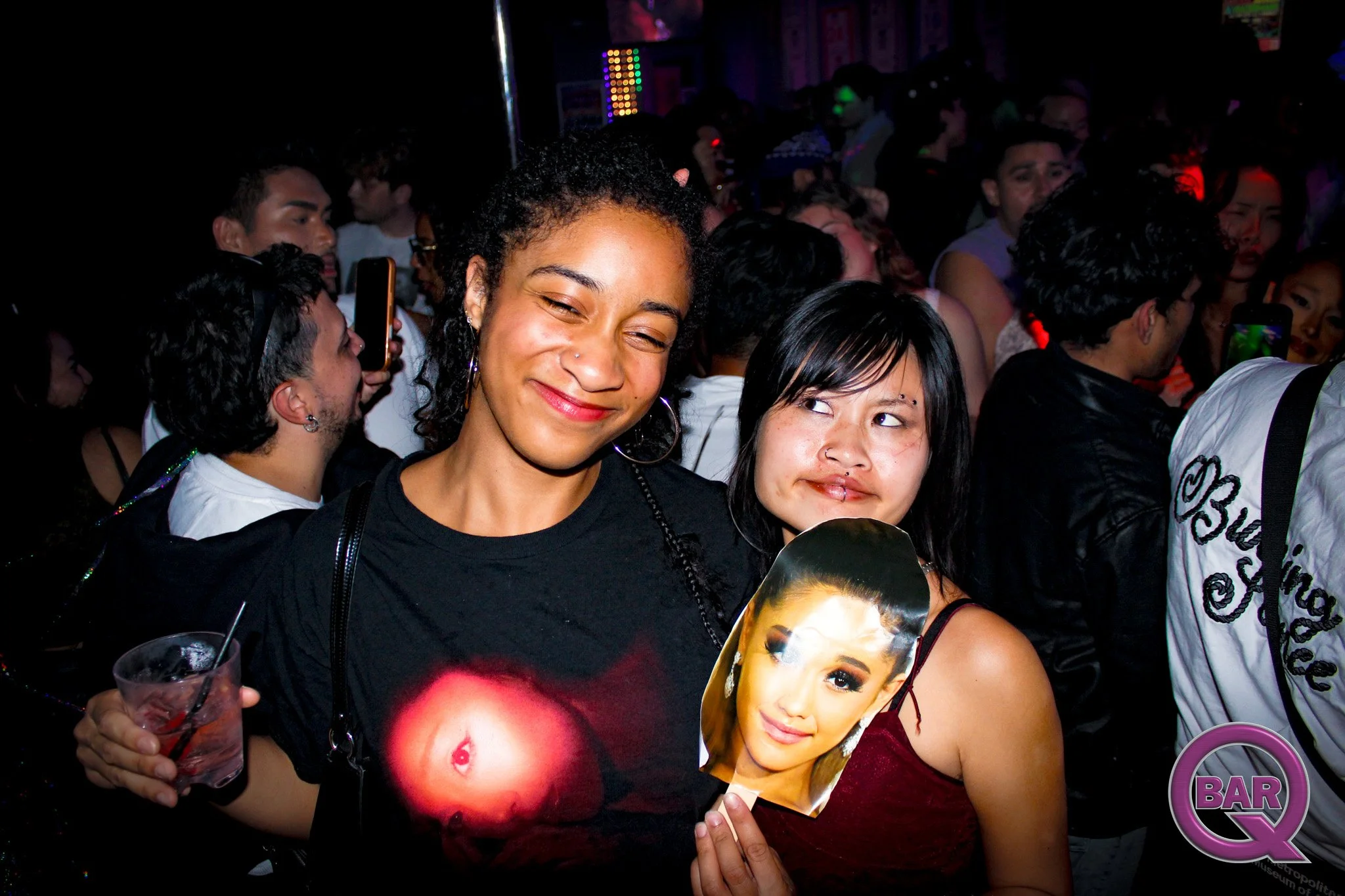 Person smiling at a crowded party, holding a fan with a woman's face. Others around are taking photos and socializing.
