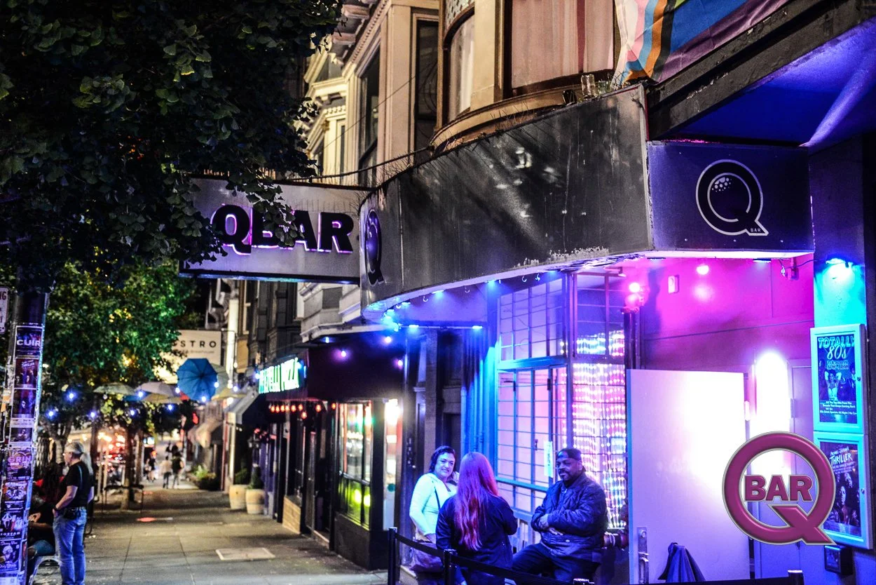 Nighttime scene of a city street with people sitting outside a bar with neon lights, illuminated signs, and posters on the wall.