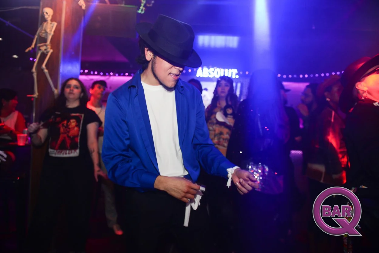 A person in a blue shirt, white undershirt, and black hat dancing in a dimly lit nightclub with purple and blue lighting, others dancing and socializing in the background, and a skeleton decoration hanging in the club.