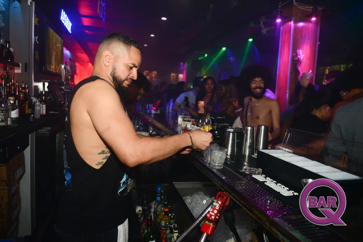 A shirtless bartender with short hair and a beard preparing drinks behind a bar in a lively nightclub with colorful lighting. Several people are dancing and enjoying themselves in the background.