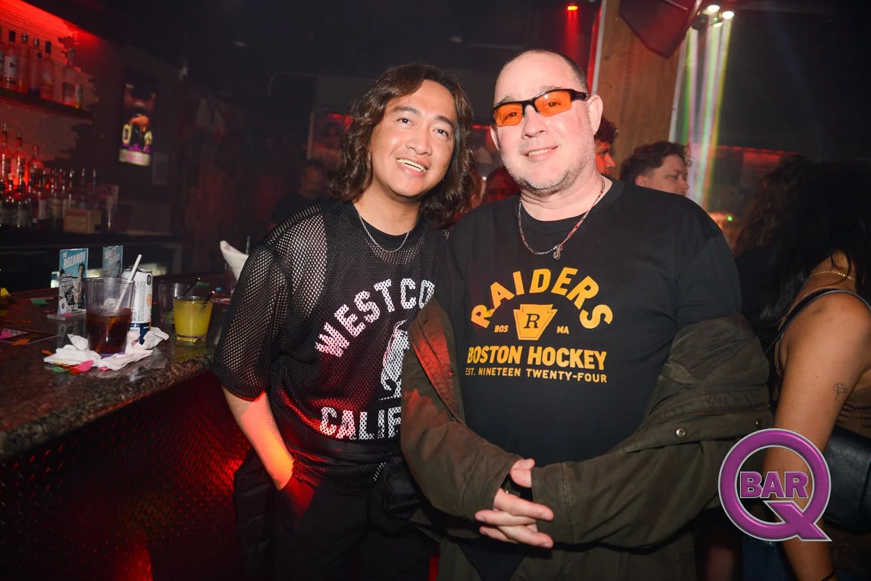 Two men posing and smiling in a bar with drinks on the counter, one wearing a mesh black West Coast California shirt, the other wearing a black Boston Hockey Raiders shirt with orange glasses, illuminated by colorful bar lights.