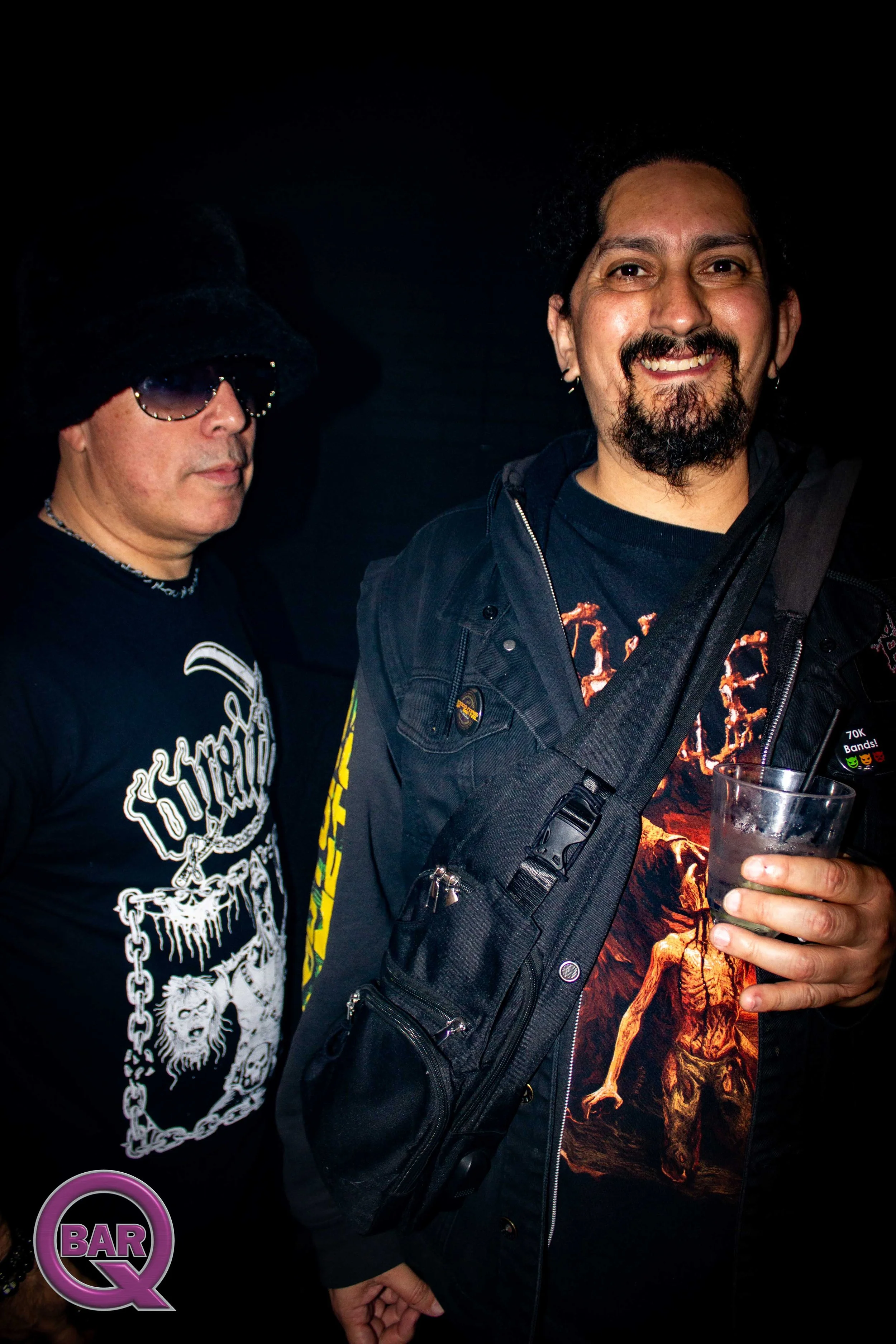 Two men standing close together in a dark setting, one wearing a black hood and sunglasses, the other smiling with a goatee and earrings, holding a glass of drink.