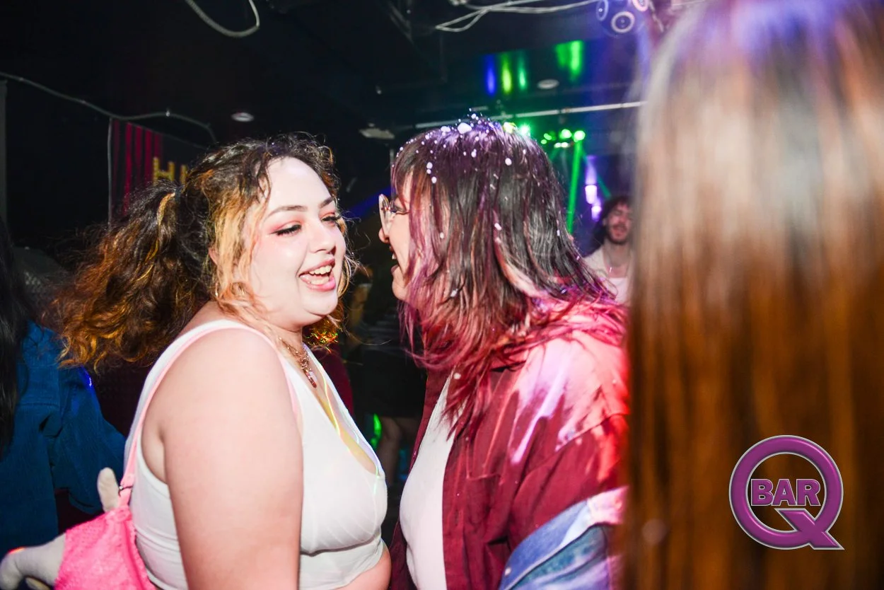 Two women smiling and enjoying themselves at a lively party or nightclub with colorful lighting, one with curly hair and the other with wet hair wearing glasses, while a third person is partially visible on the right side. A sign with the word "BARQ"