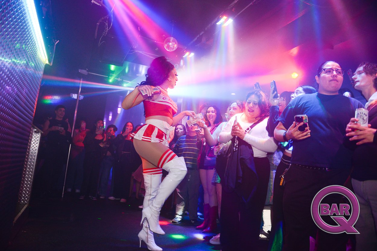 A woman dressed in a red and white cheerleader outfit with white thigh-high boots is dancing on stage at a nightclub, surrounded by a crowd of people taking photos and videos, with colorful lights and a disco ball overhead.