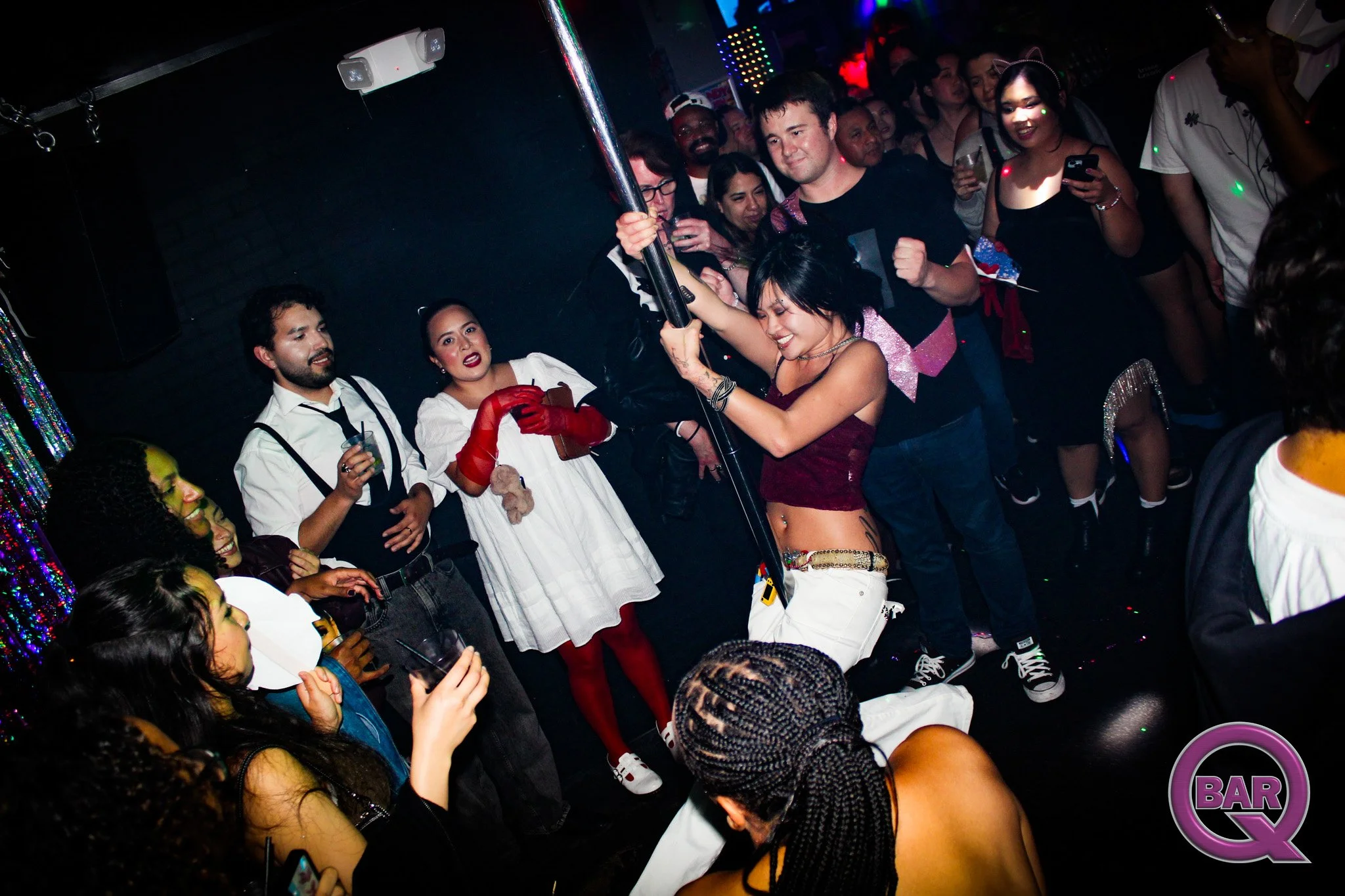 People dancing and socializing in a nightclub with colorful lighting, including a woman in lingerie holding onto a pole, surrounded by friends and partygoers.