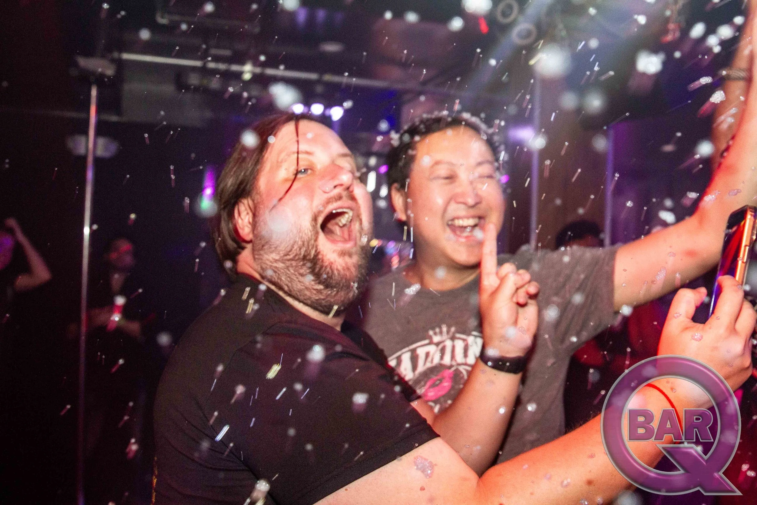 Two people celebrating at a bar with confetti falling around them, smiling and taking a selfie.