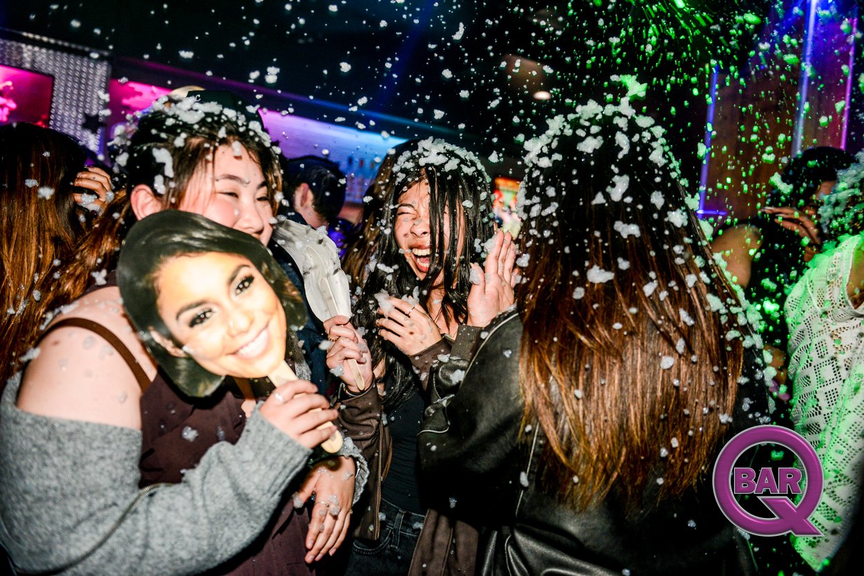 People celebrating at a bar with foam or snow falling around them, holding party masks, smiling and having fun.