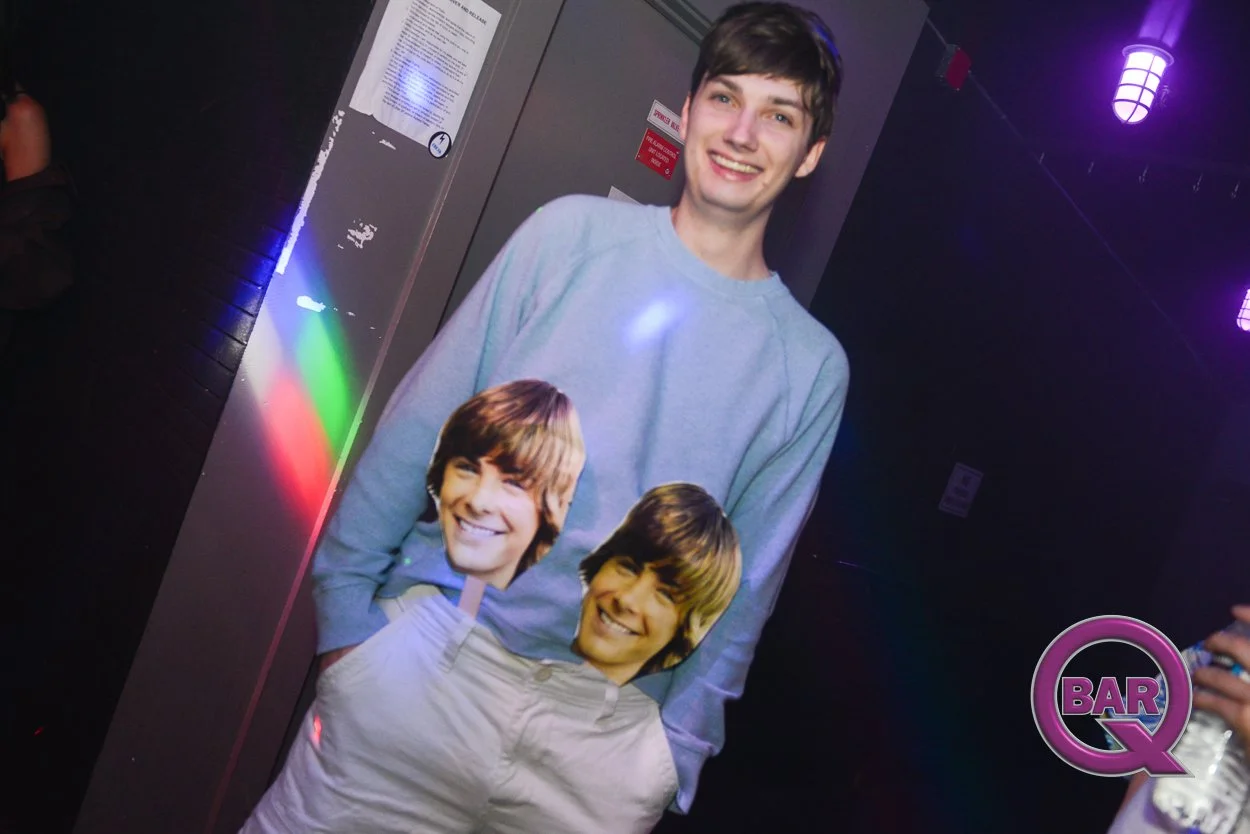 A young man standing in a dimly lit room with purple and green lights, wearing a sweatshirt with multiple images of a man's smiling face on it, and holding a drink in his hand.