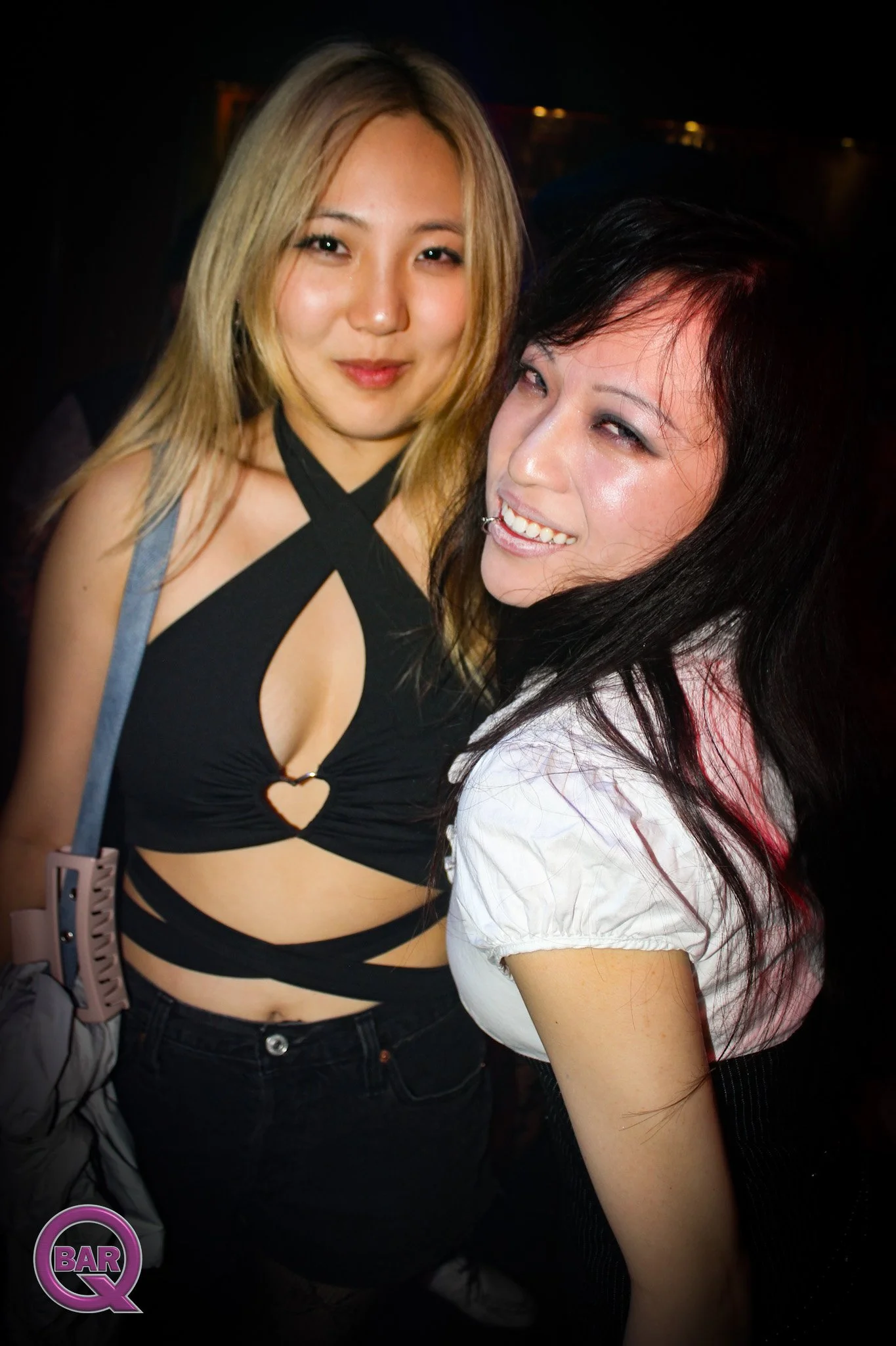 Two women smiling and posing for a photo in a dimly lit environment, possibly a nightclub or bar, with one woman wearing a revealing black top with cutouts and the other in a white shirt.