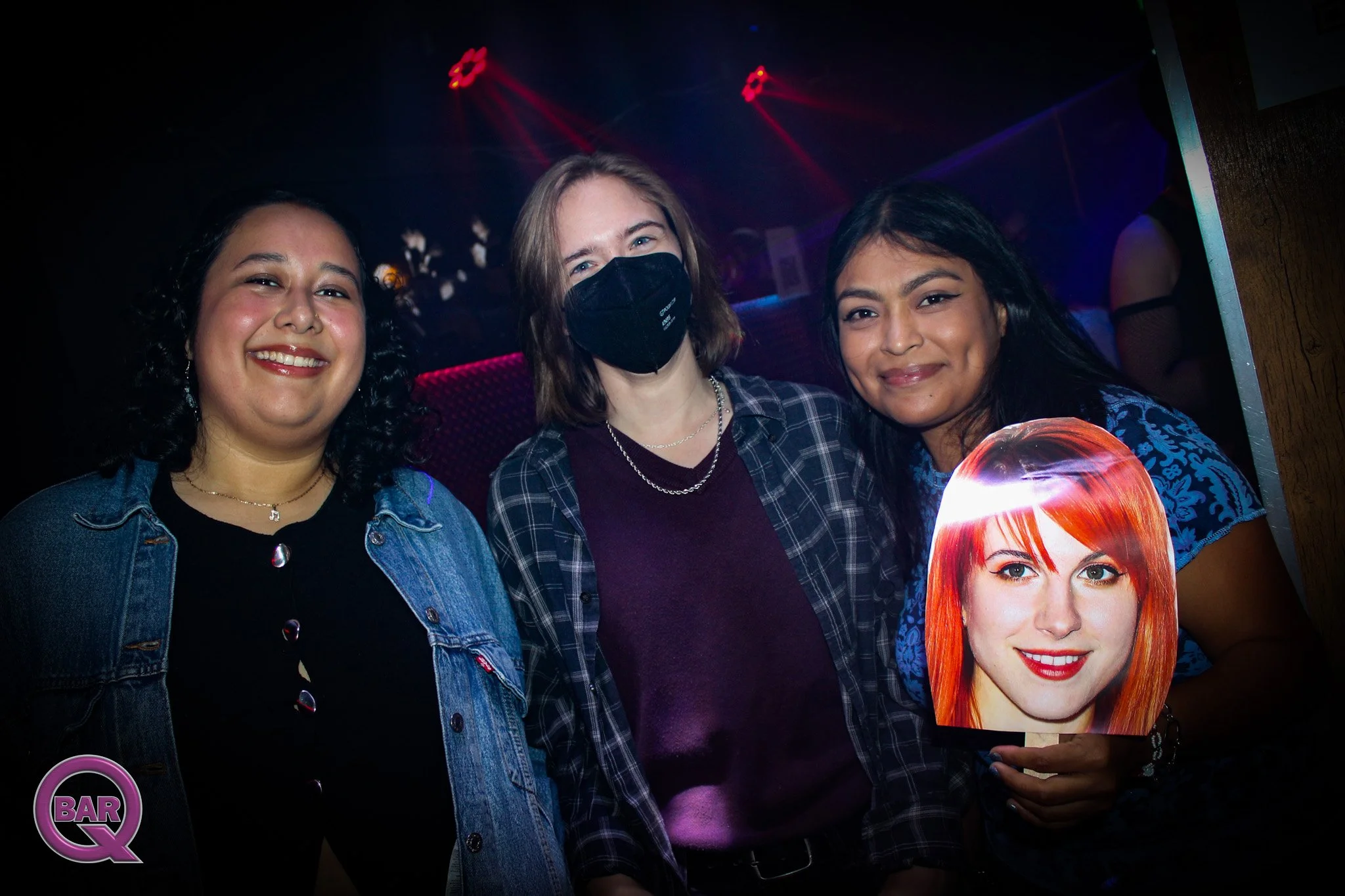 Three women standing together at a nightclub with colored lights in the background. The woman on the left has dark curly hair and is smiling. The woman in the middle has light brown hair, is wearing a black face mask and a plaid shirt. The woman on t
