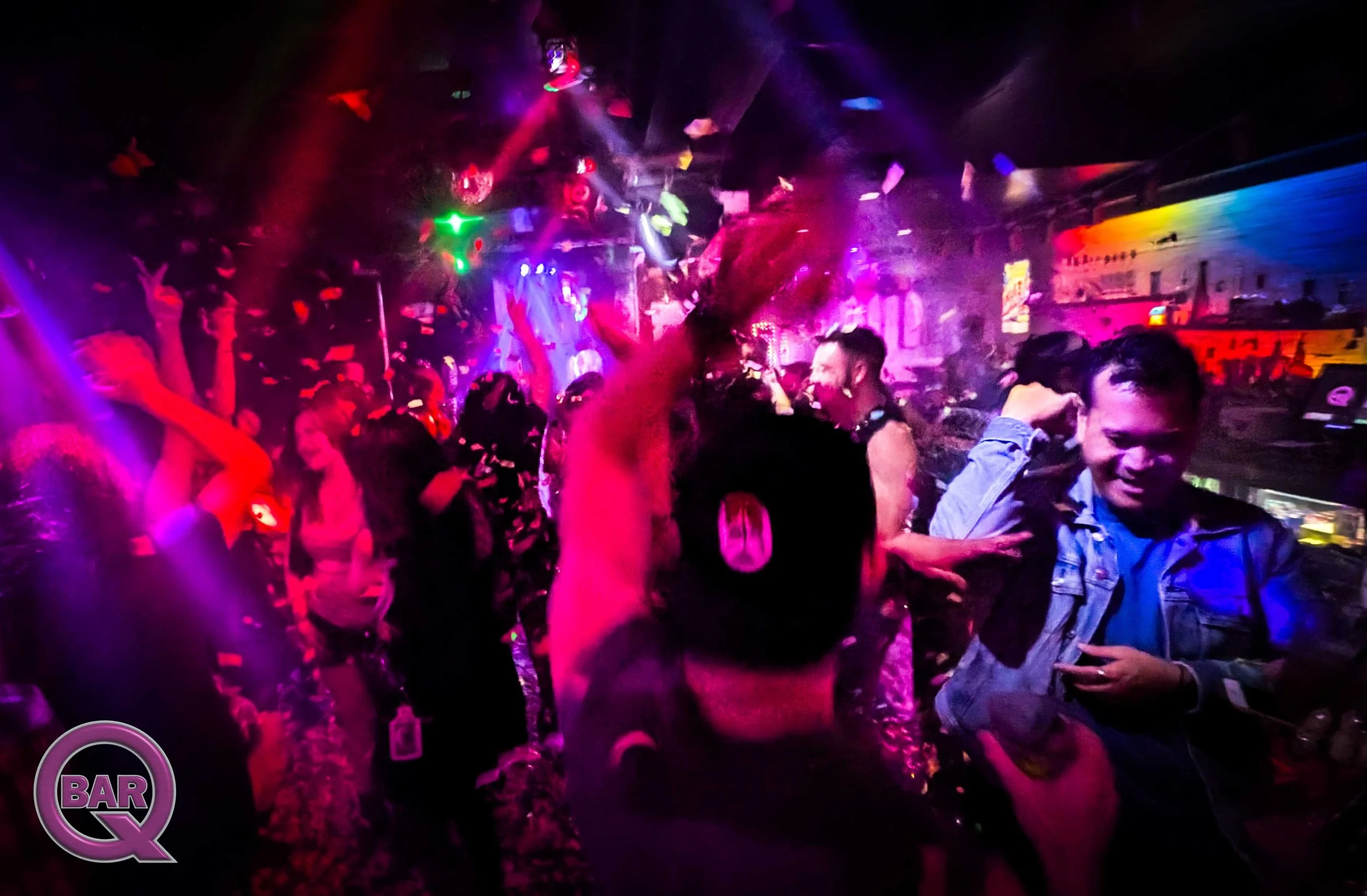 People dancing and socializing in a nightclub with colorful lighting and a bar in the background.