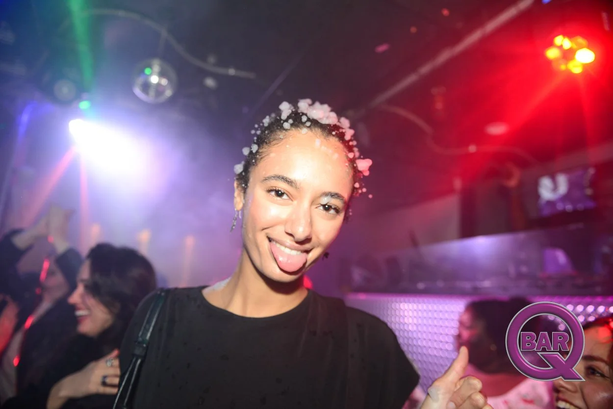 Young woman with wet hair and foam on her head at a nightclub, smiling and sticking out her tongue, with colorful lights and other people in the background.
