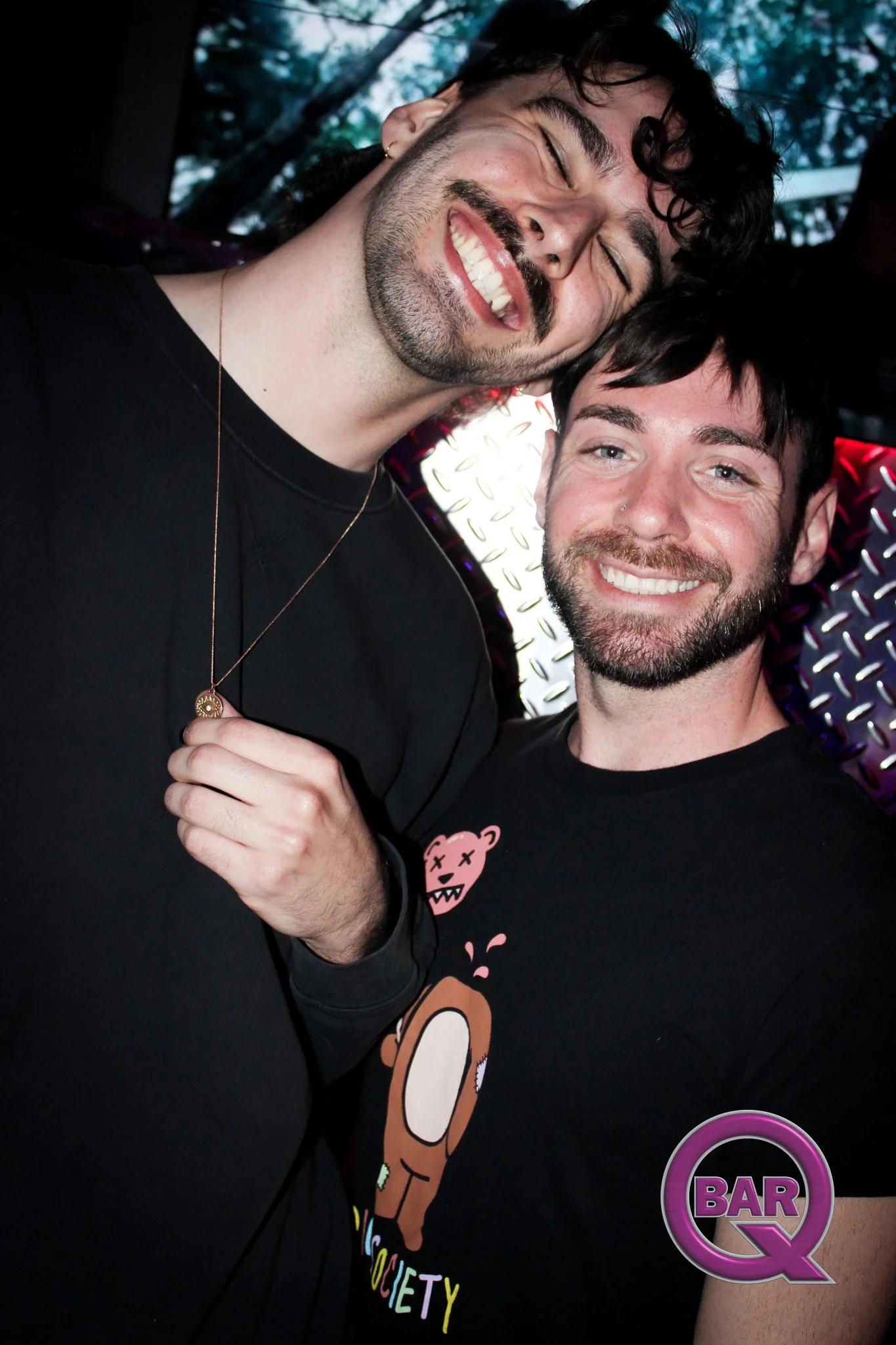 Two men smiling and enjoying themselves at a bar or club, with a vibrant, patterned background and a neon sign that reads 'BAR Q'.