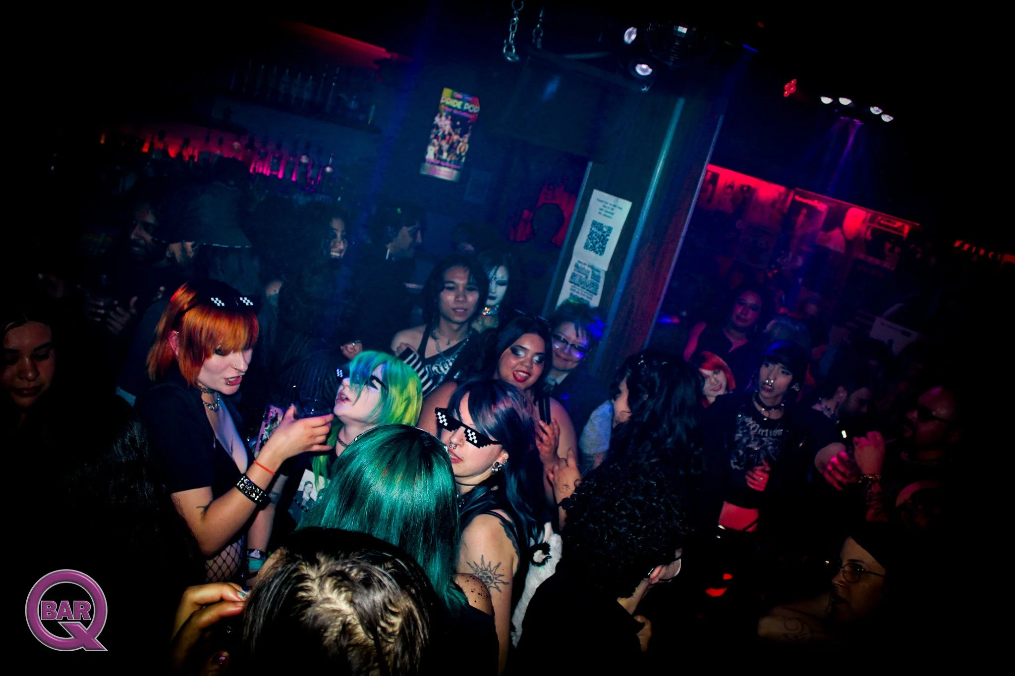 Crowd of people at a nightclub with colorful lighting, some wearing sunglasses and with bright, colorful hair.