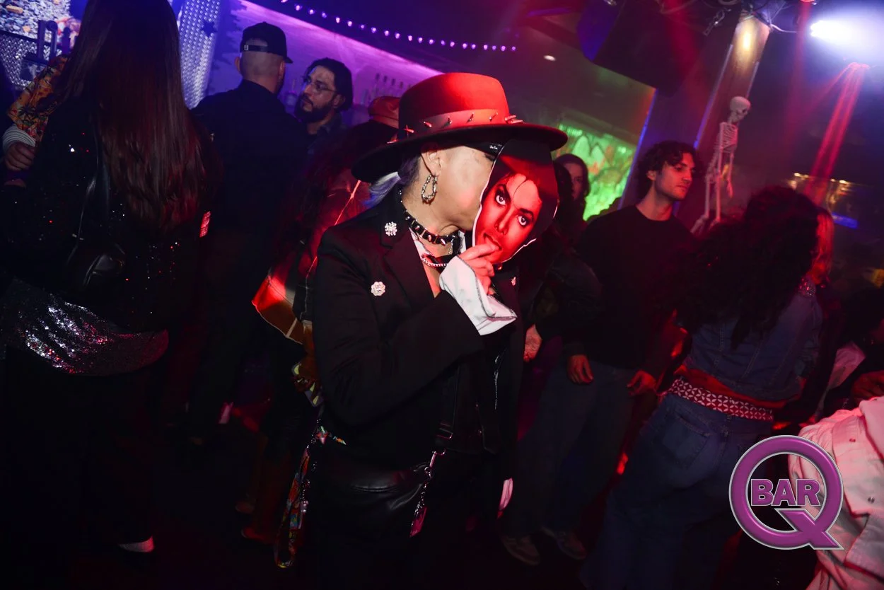 People dancing and socializing in a dark, colorful nightclub with neon lights. A woman in the center wears a red hat and a mask with a woman's face on it, holding a finger to her lips. The background features a skeleton decoration and a purple and gr