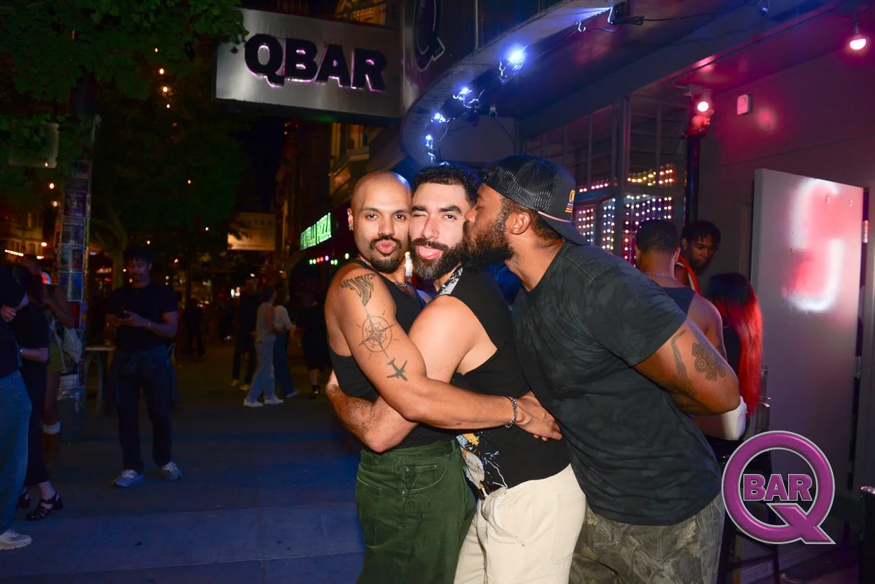 Three men embracing and posing for the camera outside a bar at night, with other people in the background; one is kissing another on the cheek, and the scene is illuminated by colorful lights.