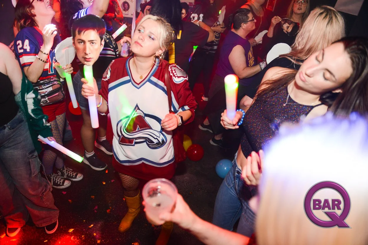 People dancing at a party or nightclub holding glow sticks, with some balloons on the floor, in a lively setting with colorful lighting and a sign that says 'Bar Q'.