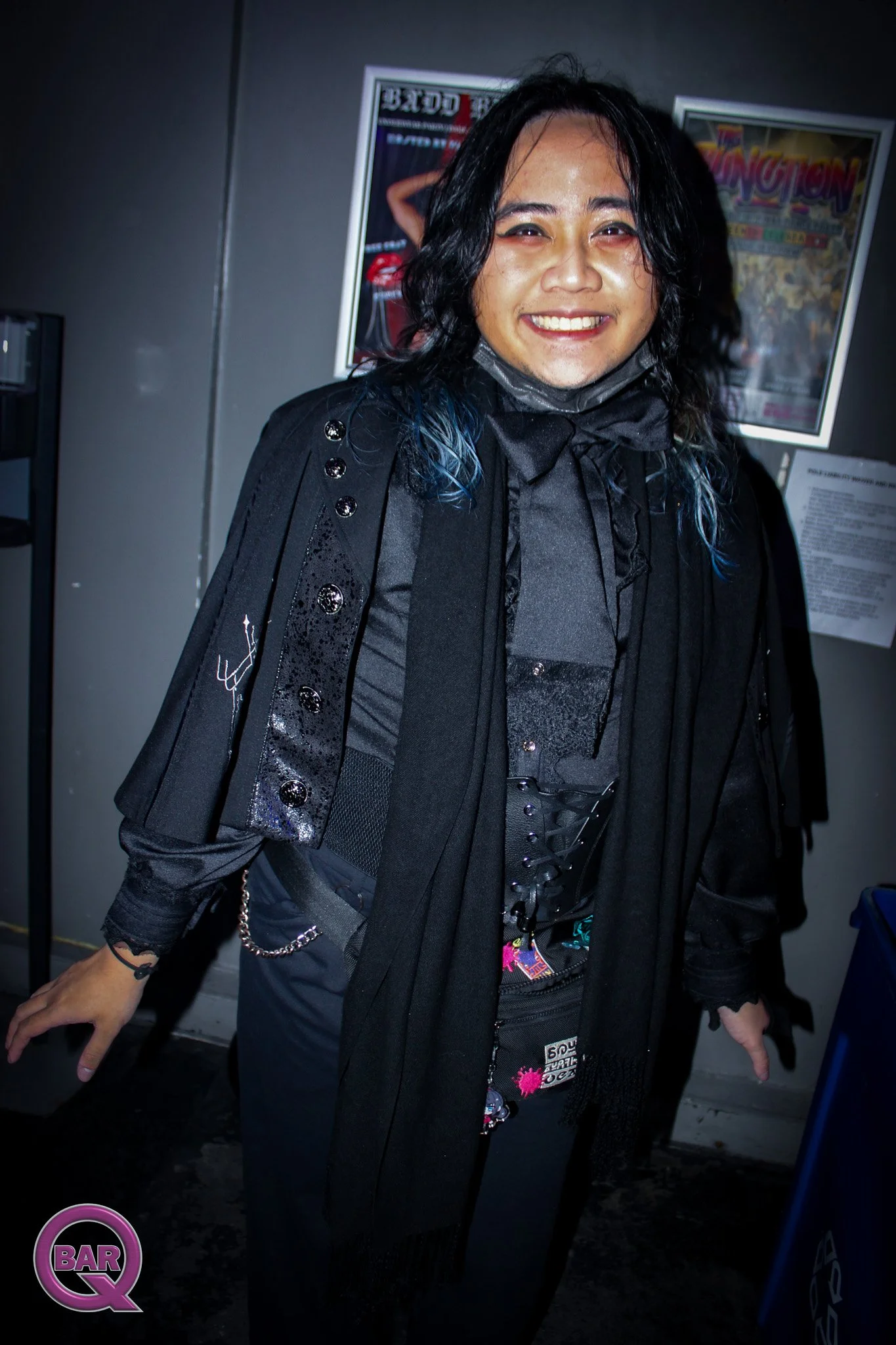 Person with long black wavy hair, wearing black clothing with silver details, smiling in front of a dark wall with posters.