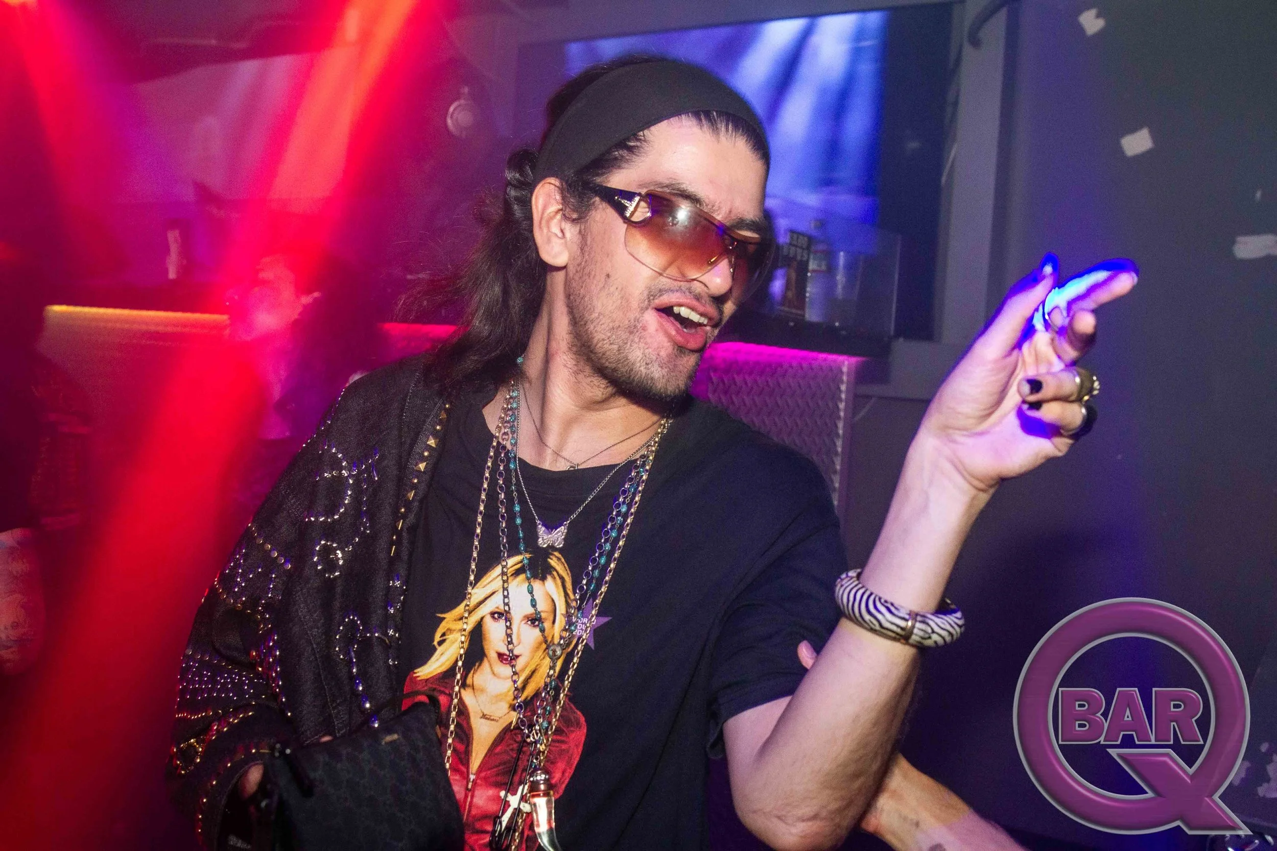 A person with long dark hair, wearing a black headband, sunglasses, and layered jewelry, is dancing or enjoying music in a nightclub with purple and pink lighting.