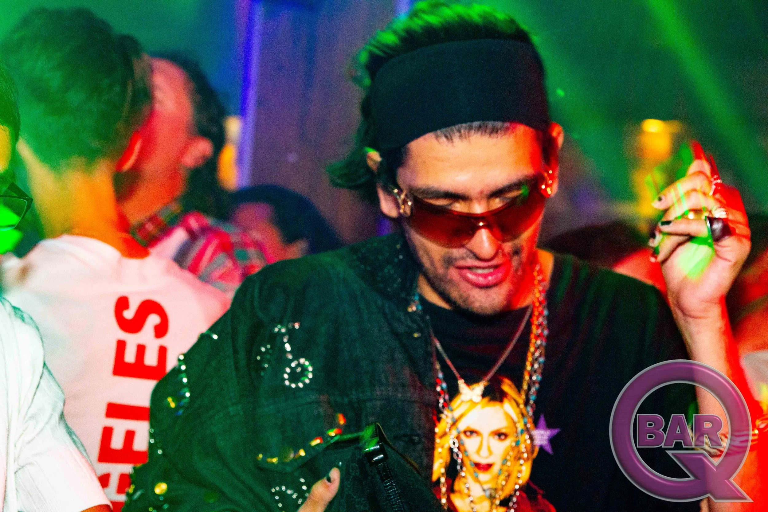 A person with green-styled hair, wearing sunglasses, a black headband, and jewelry, at a lively event with colorful lighting, surrounded by other people, including someone holding a large cup and a woman on a poster.