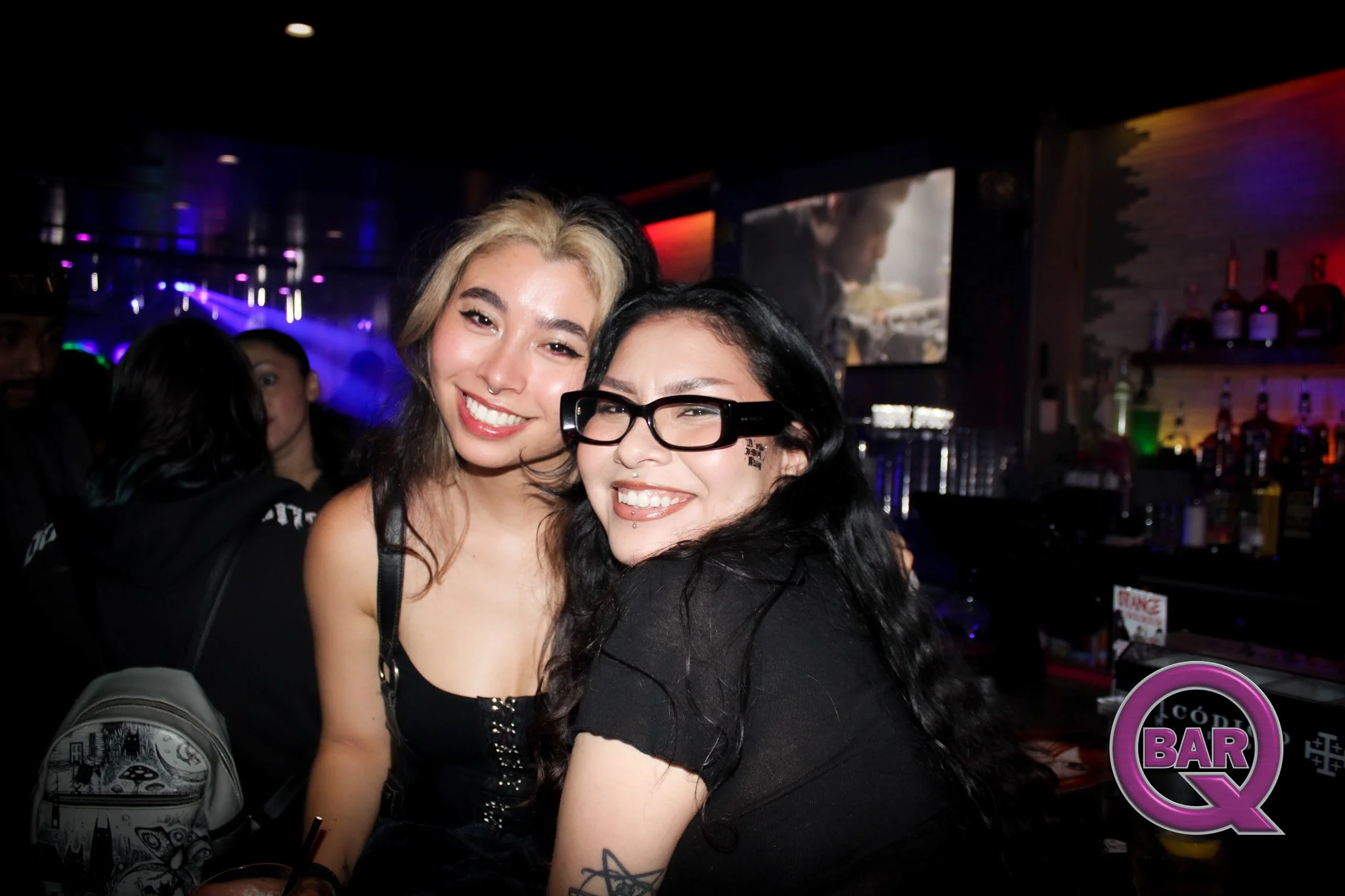 Two women smiling and hugging in a bar with colorful lighting and a video screen in the background.