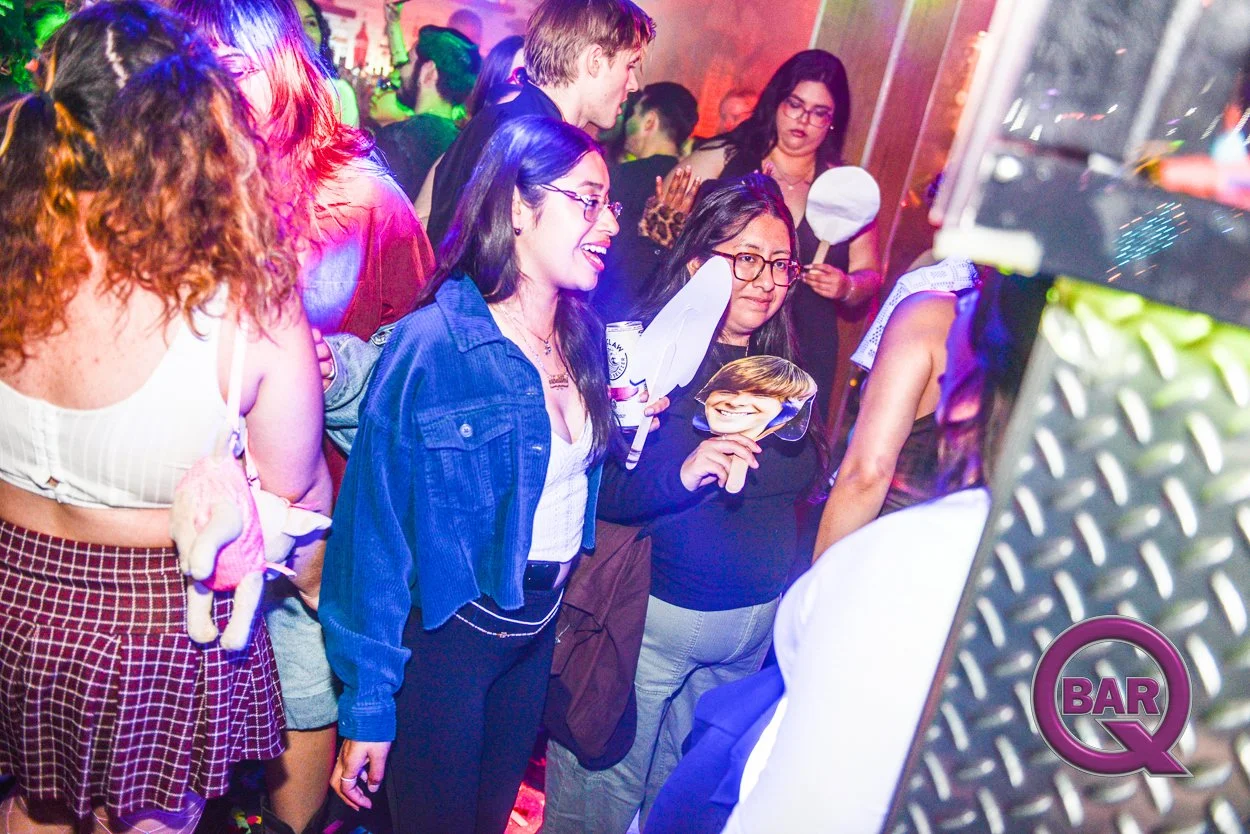 Group of people at a bar or nightclub, some wearing costumes, enjoying music and socializing with colorful lighting and a 'Bar Q' sign in the foreground.
