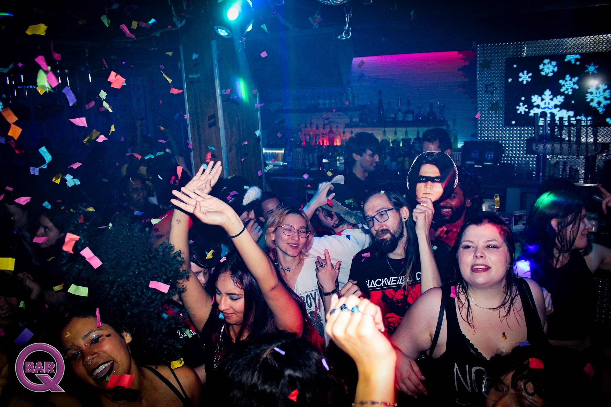 A crowded nightclub with people dancing and celebrating, colorful confetti falling from above, and a bar in the background with snowflake decorations and vibrant lighting.