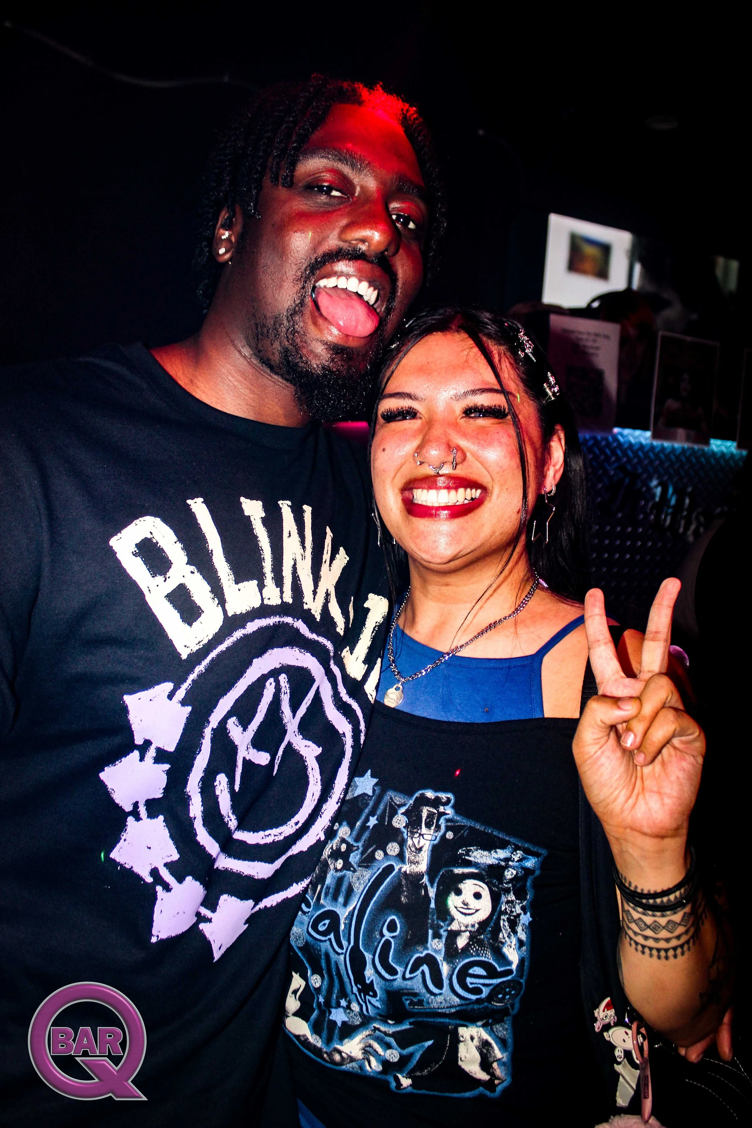 Two people smiling and posing for a photo at a bar or club. The man on the left has dark skin, curly hair, and is sticking out his tongue. The woman on the right has light skin, long dark hair with clips, multiple facial piercings, and is showing a p