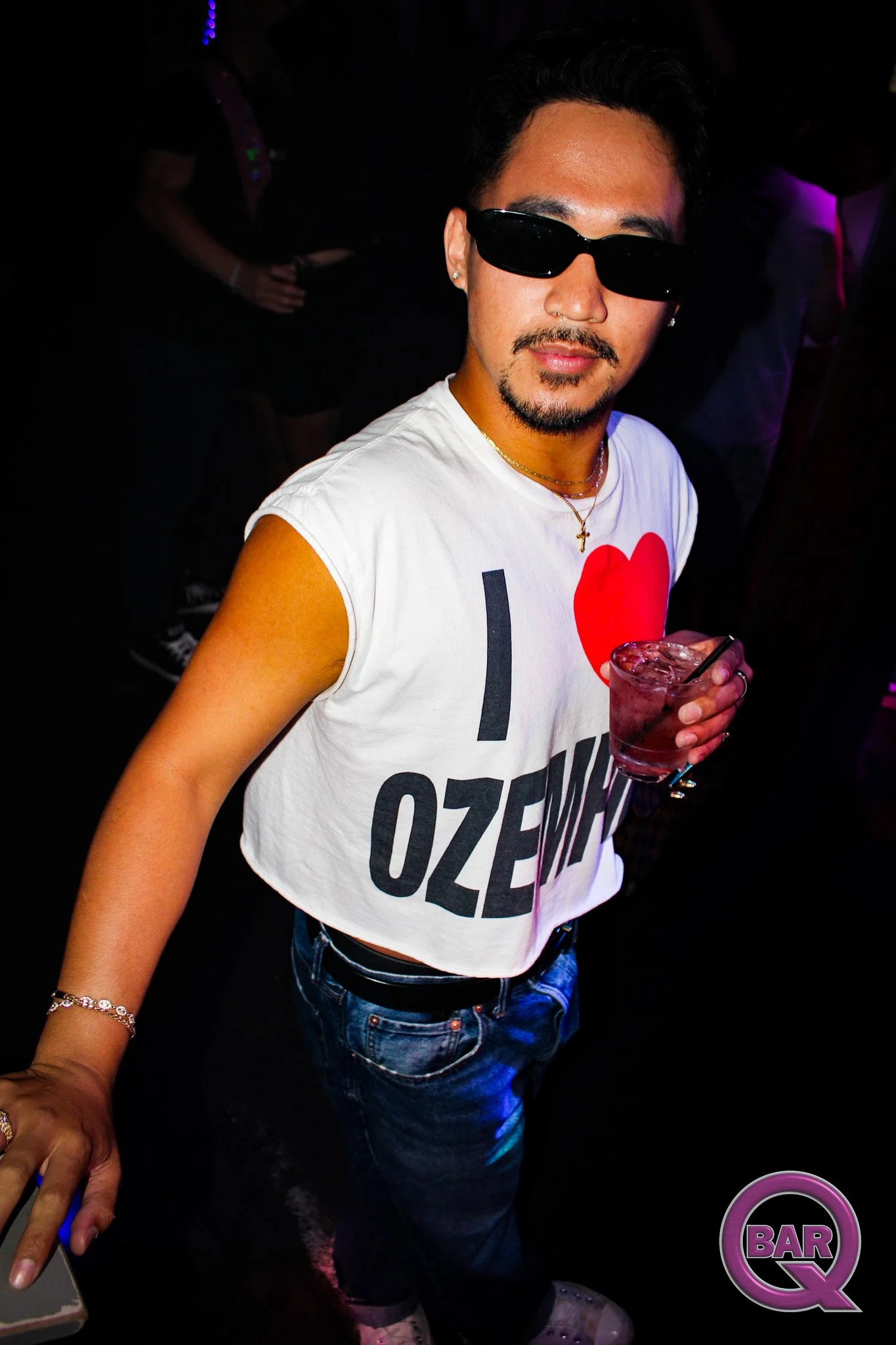 A man wearing sunglasses and a white sleeveless t-shirt with a red heart graphic and text, holding a drink in a nightclub with colorful lights and a purple 'Bar Q' sign in the bottom right corner.