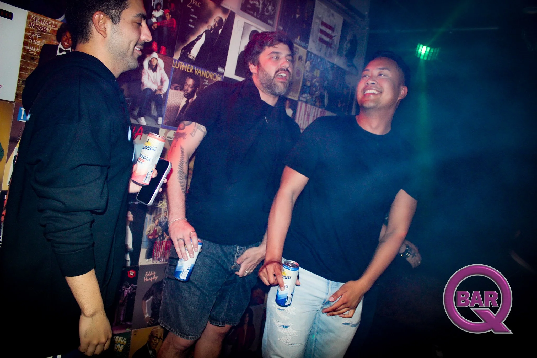 Three men are standing at a bar, holding cans of canned iced tea, and smiling. There are movie posters and a neon sign behind them.