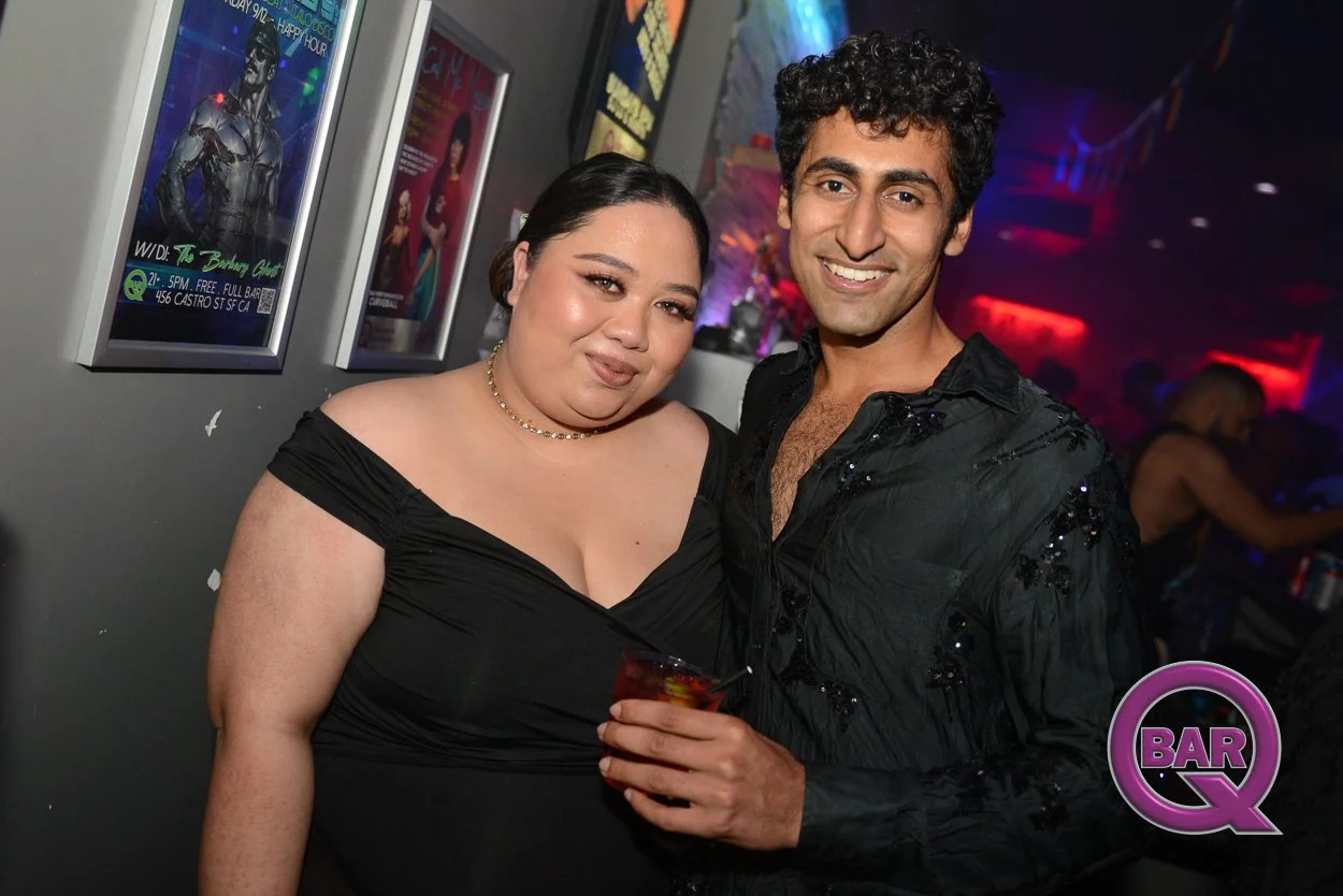 Two people, a woman with dark hair and a man with curly dark hair, smiling and standing together in a nightclub or bar with colorful lights in the background. The woman is wearing a black off-shoulder dress with a necklace and the man is wearing a bl