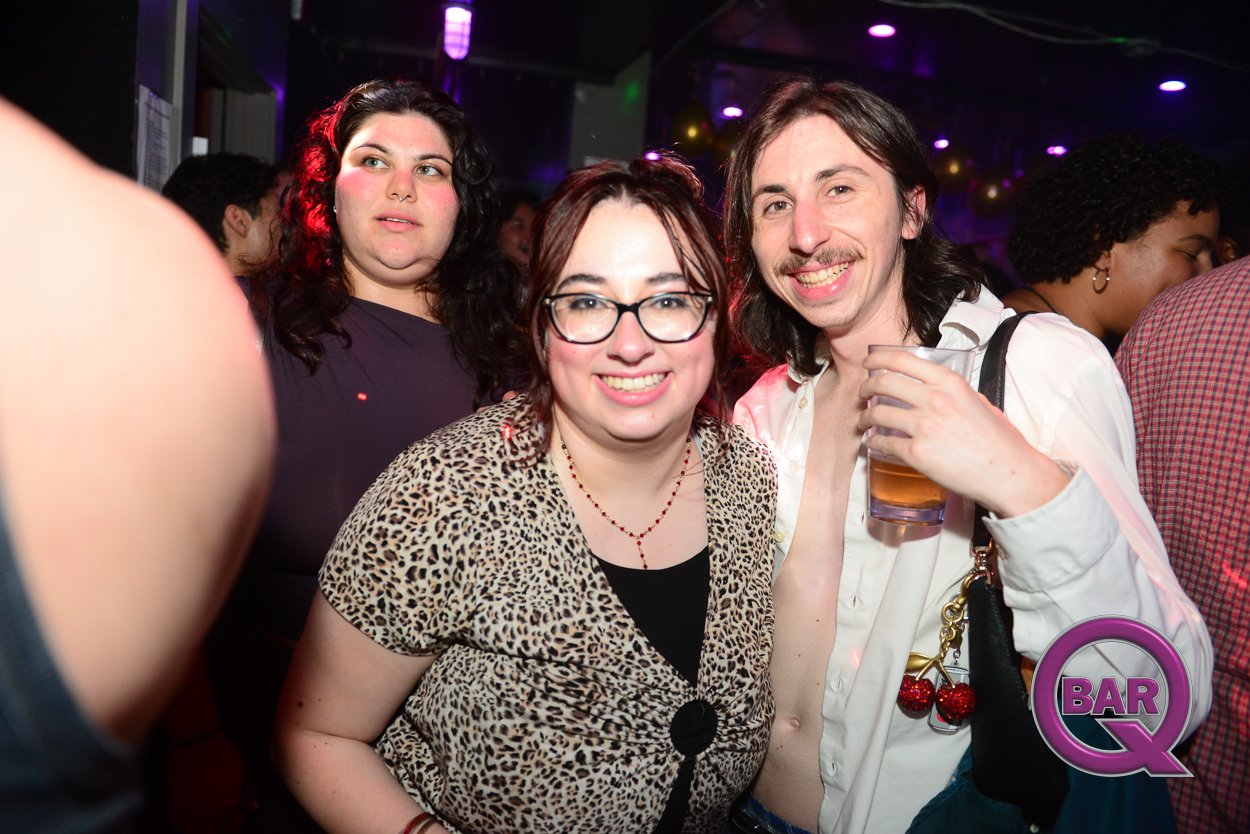 Group of people at a party or bar with colorful lighting, including two women and one man smiling at the camera. The man has long hair and is holding a drink, wearing a white shirt with cherries on his purse. The woman in the middle has glasses, dark