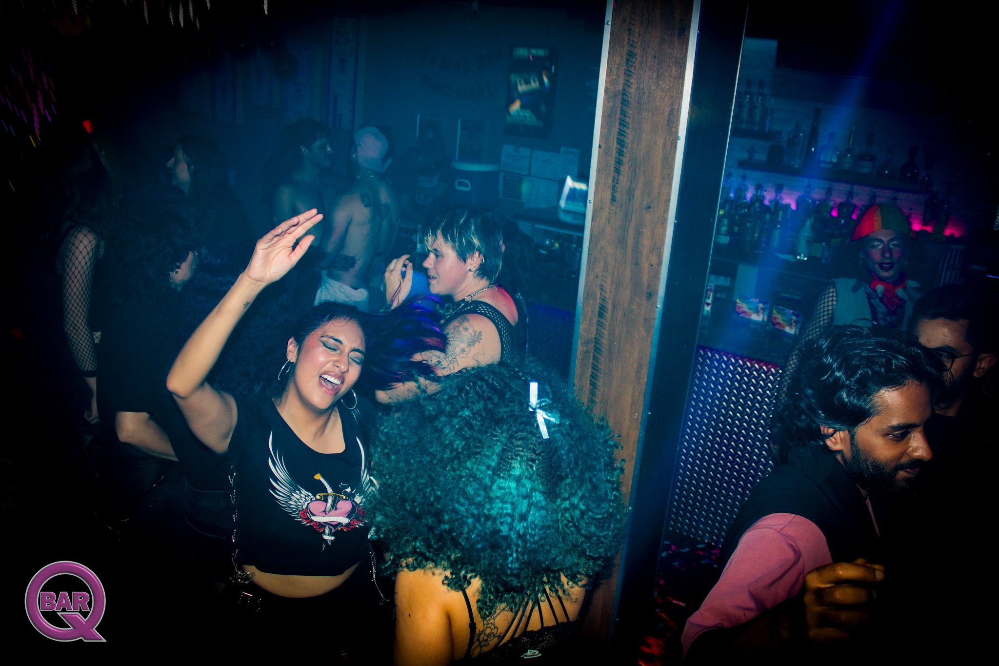 Women dancing at a nightclub with colorful lighting, bar area in the background, and some people wearing fishnet clothing and costumes.